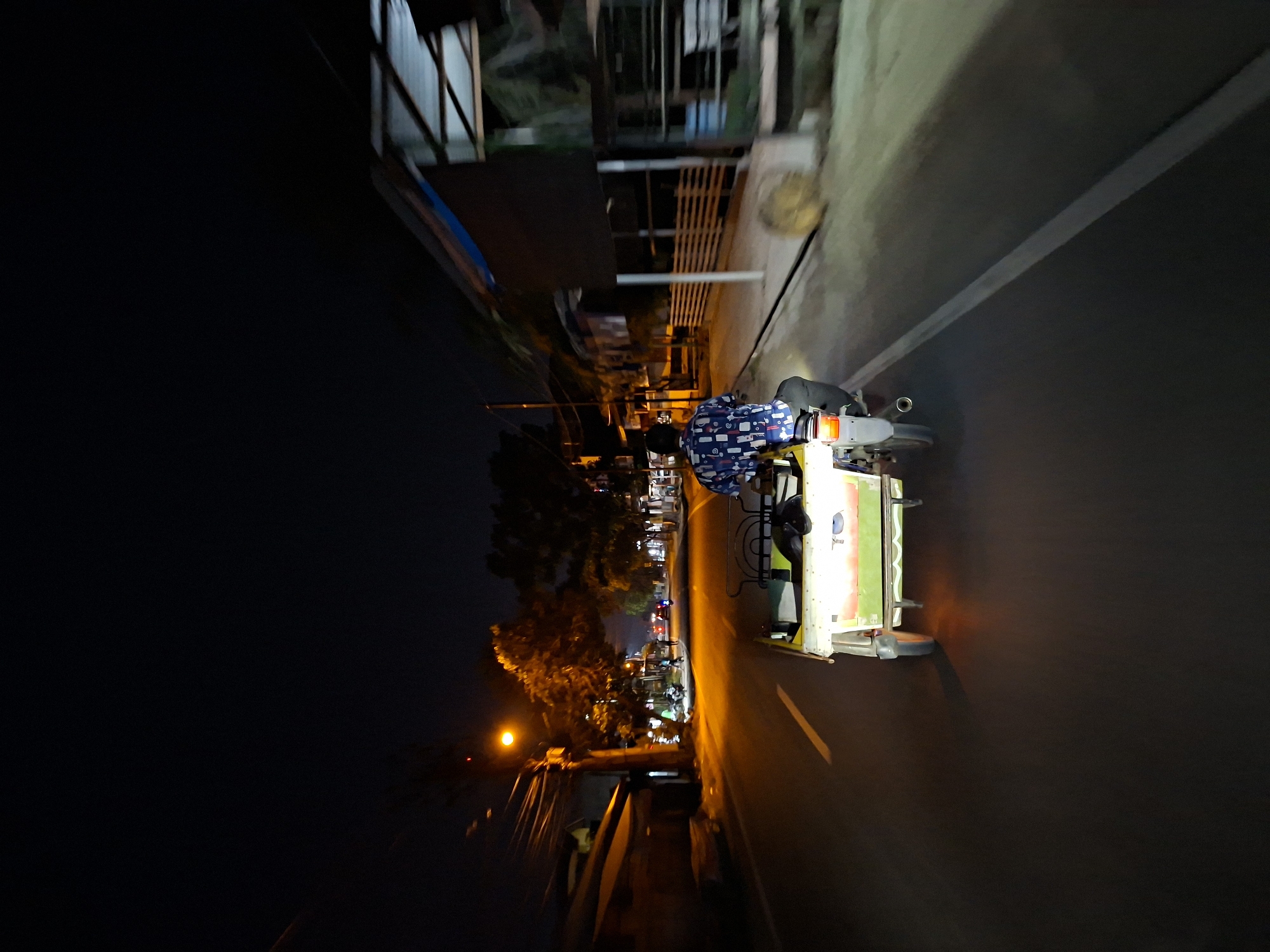 An overhead, slightly angled shot captures a motor-rickshaw traveling along a paved road at night in Lhokseumawe, Indonesia. The image is significantly rotated clockwise.
The primary subject is a motor-rickshaw, also known as a 'becak motor', which consists of a motorcycle attached to a light-colored sidecar. The sidecar features a light green top or canopy and white lower sections. A person, presumably the driver, is seated on the motorcycle, wearing a blue shirt with a white pattern. The red taillight of the motorcycle is illuminated.
The vehicle is in motion, indicated by the motion blur present in the background and on the road surface, which features a faint white dividing line. Streetlights illuminate parts of the scene with warm, orange-yellow light, casting a glow on the road and nearby structures. On the left side of the road (when viewed correctly, not rotated), several trees and blurred building structures are visible, also partially lit. The right side of the road shows more indistinct background elements and structures. The sky is dark, emphasizing the artificial illumination. No legible text is visible in the image.