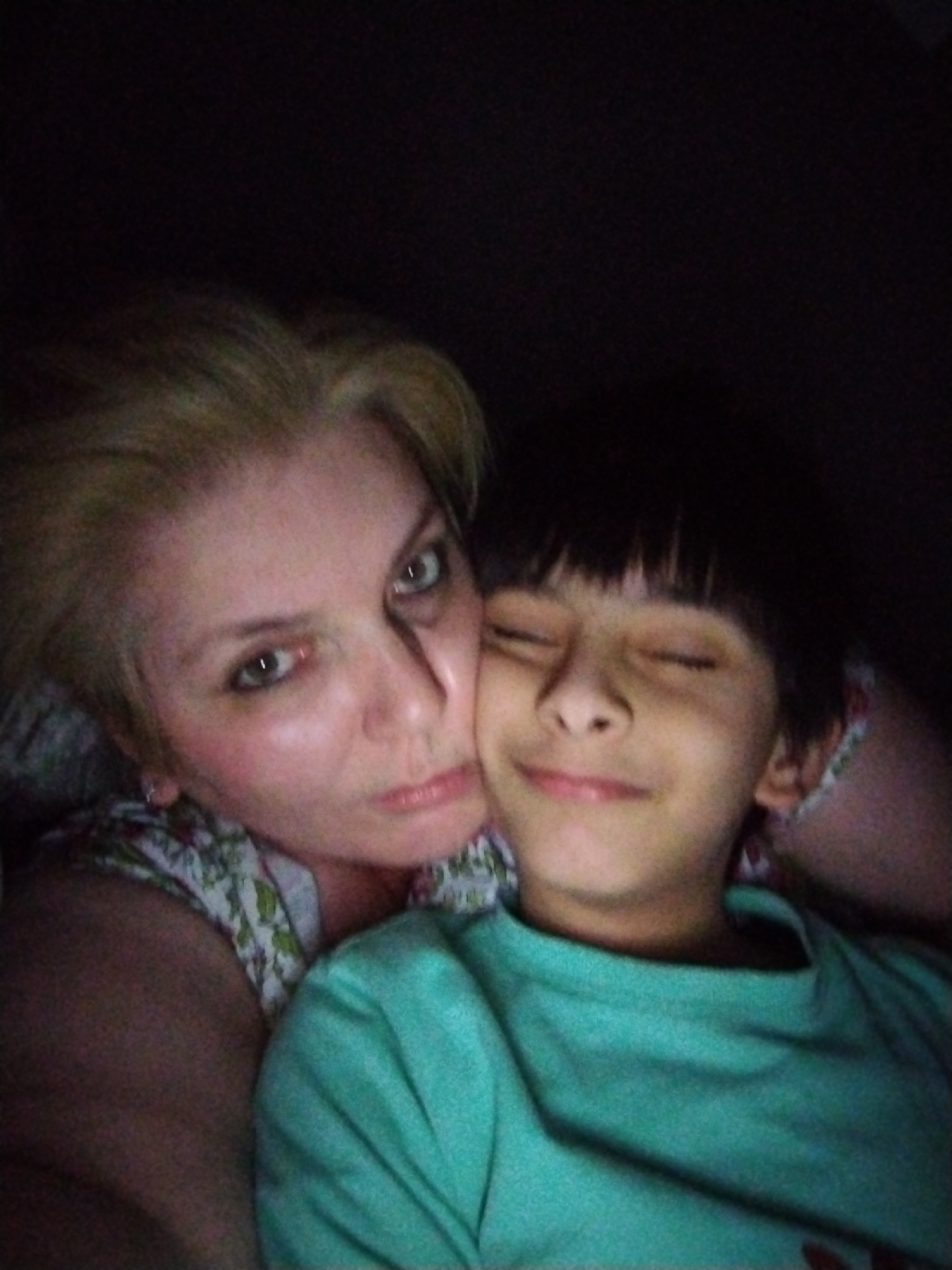 A low-light, medium-close-up image depicts an adult woman and a child in what appears to be an indoor setting. The woman, positioned on the left and partially obscured, has short, light-colored hair and light skin. Her eyes are open, focused towards the camera, displaying a direct gaze. She has a subtle, neutral to slightly serious expression. Her attire includes a light-colored top with a floral or patterned design visible on her shoulder and chest. Her face is in close proximity to the child's head. The child, positioned on the right, has dark, straight hair with bangs. Their eyes are closed, and a gentle smile is present on their face, indicating contentment or peacefulness. The child is wearing a plain teal or turquoise long-sleeved shirt and appears to be leaning against the woman. The background is predominantly dark, suggesting a dimly lit environment with frontal illumination on the subjects' faces.