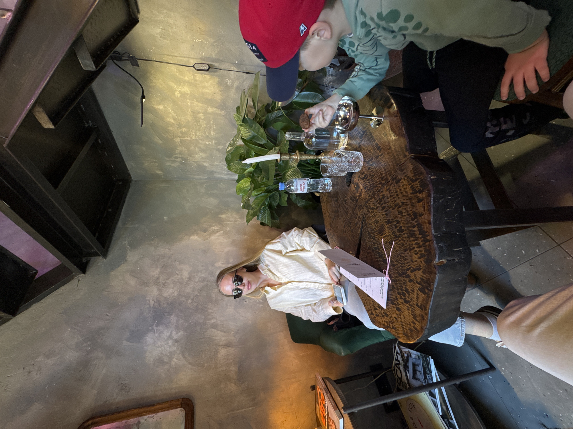 A woman in sunglasses and a light-colored shirt is seated at a uniquely shaped wooden table. A young child wearing a red cap and a green patterned sweatshirt is standing at the same table, interacting with items such as wine glasses, bottles, and a candle. In the foreground, a portion of a magazine rack is visible. The location is indoors, with a gray wall and structural steel elements overhead. The table is decorated with greenery and bottles. The woman is holding a menu. The scene takes place in Krasnaya Polyana, Russia.