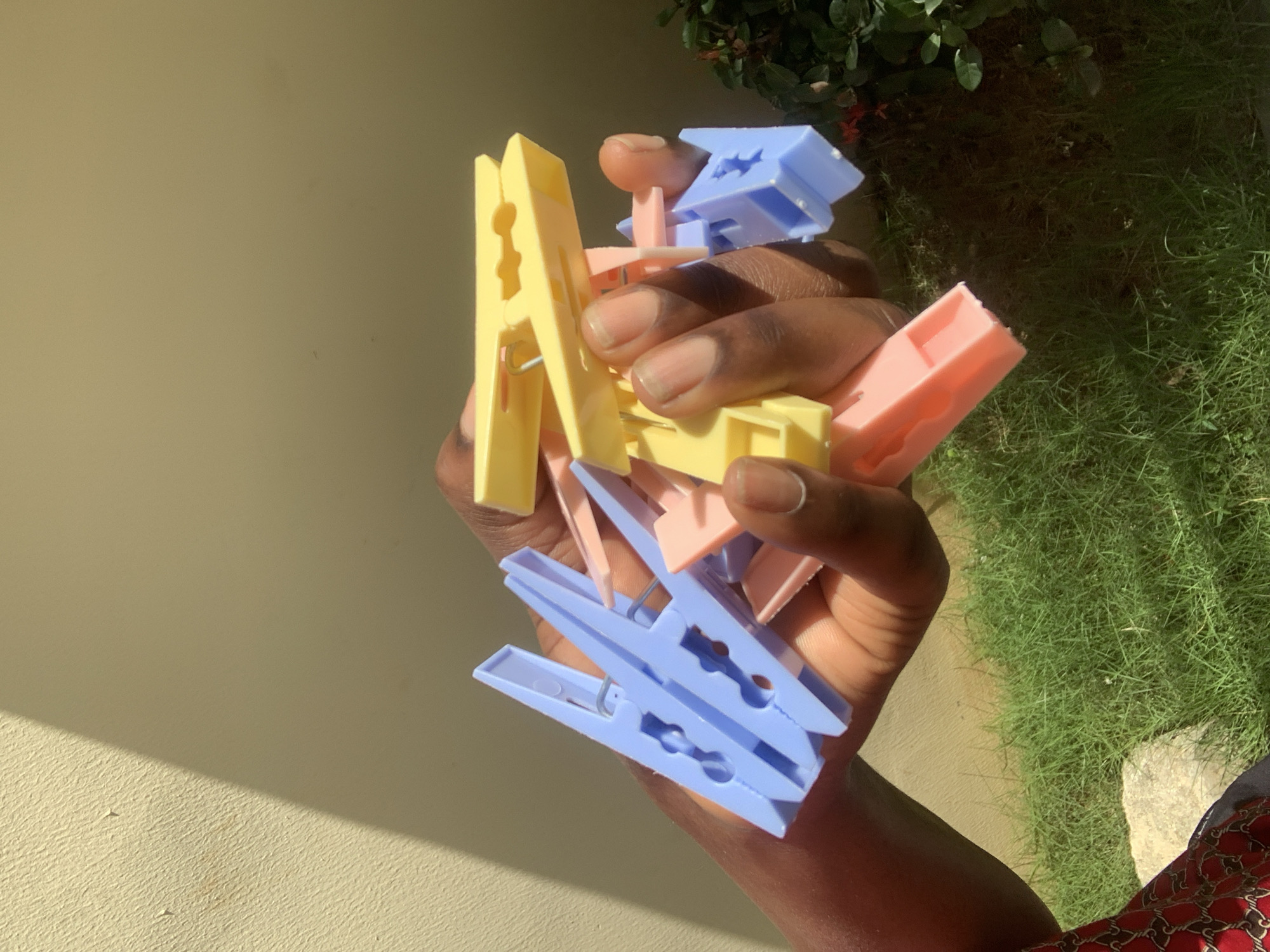 In the city of Abuja, Nigeria, a person's hand holds a cluster of colorful clothespins against a light-colored wall and a patch of green grass. The clothespins are a mix of blue, yellow, and pink. Sunlight casts a shadow on the wall, creating a diagonal line across the scene. The background also includes some green foliage. The image is a close-up shot, focusing on the hand and the clothespins.