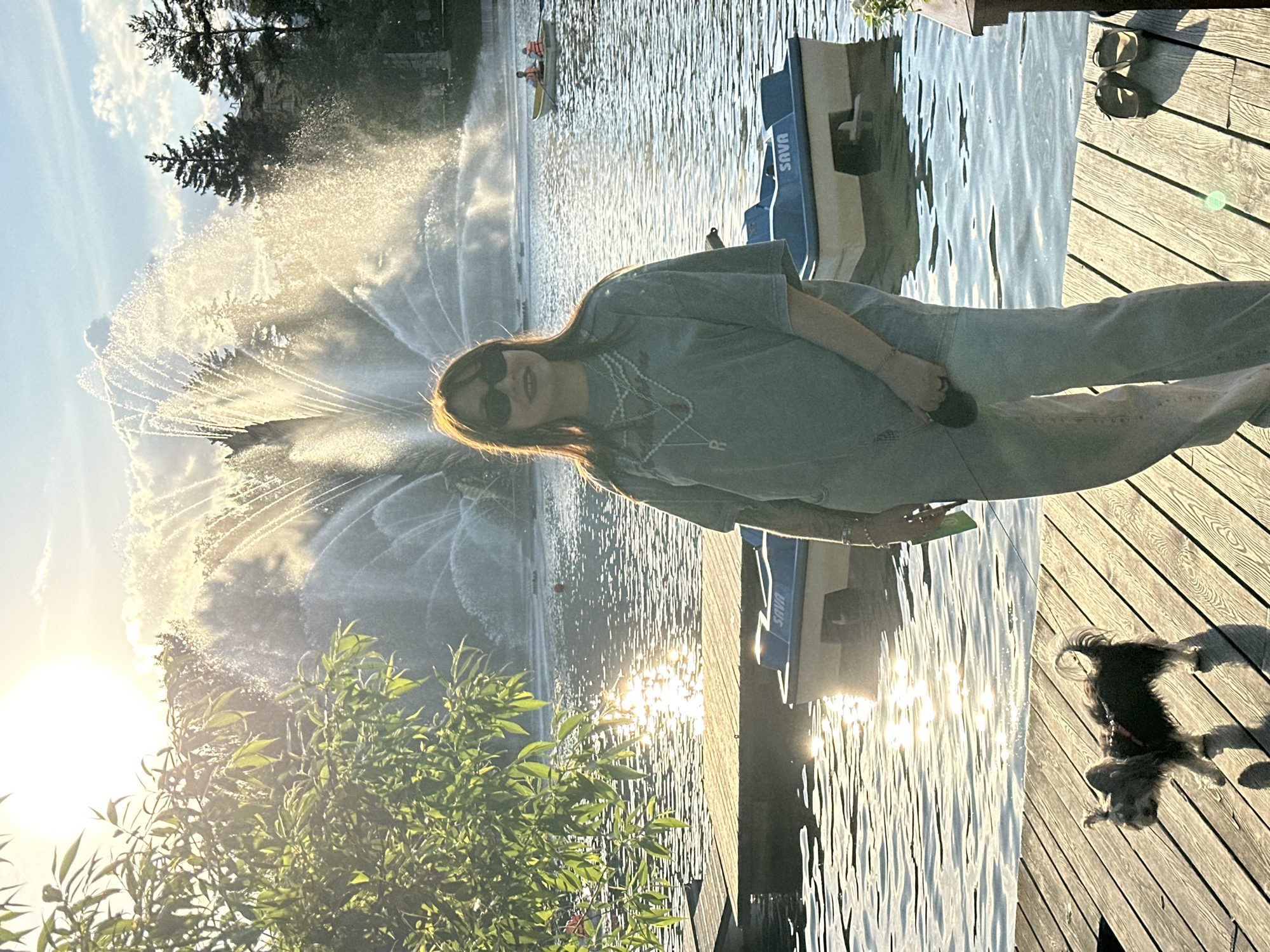 This image depicts a sunny, late afternoon scene at a recreational lake or riverfront in Moscow, Russia. A young woman stands on a wooden dock on the right side of the frame, looking towards the viewer. She is wearing dark sunglasses, a light-colored t-shirt with a beaded necklace, and jeans. The setting sun, located in the lower left, casts a warm, golden glow across the scene, creating strong highlights and a sunburst effect on the rippling water.
In the mid-ground, a large water fountain actively sprays, its streams catching the sunlight and appearing luminous. Further out on the water, at least one person can be seen in a small rowboat or kayak. Several blue-and-white pedal boats or paddle boats are docked near the woman, and the word "SAVA" is clearly visible on the side of one of them.
On the wooden deck in the lower right, a small, dark-furred dog is lying down, casting a distinct shadow. Green foliage is present in the foreground on the left, partially silhouetted by the intense sunlight. Beyond the water and fountain, evergreen trees line the distant shore beneath a sky with scattered clouds. The scene conveys a relaxed and pleasant outdoor atmosphere.