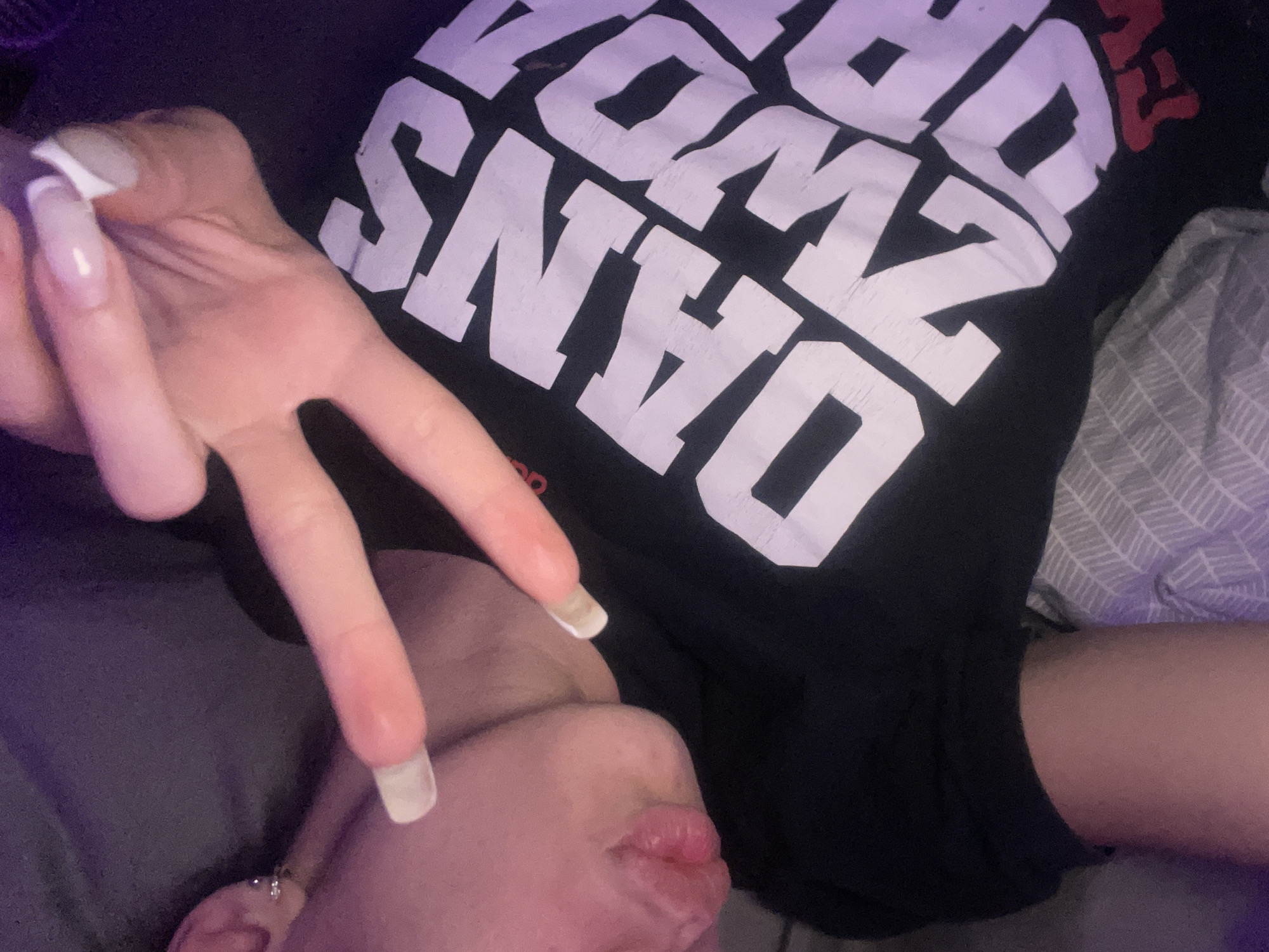 The image shows a close-up of a person's face and chest, with a hand extended in a peace sign. The person is wearing a black t-shirt with large white lettering. The lighting in the image is dim, with a purplish hue. There is no visible indication of the location in Totleben, Bulgaria.