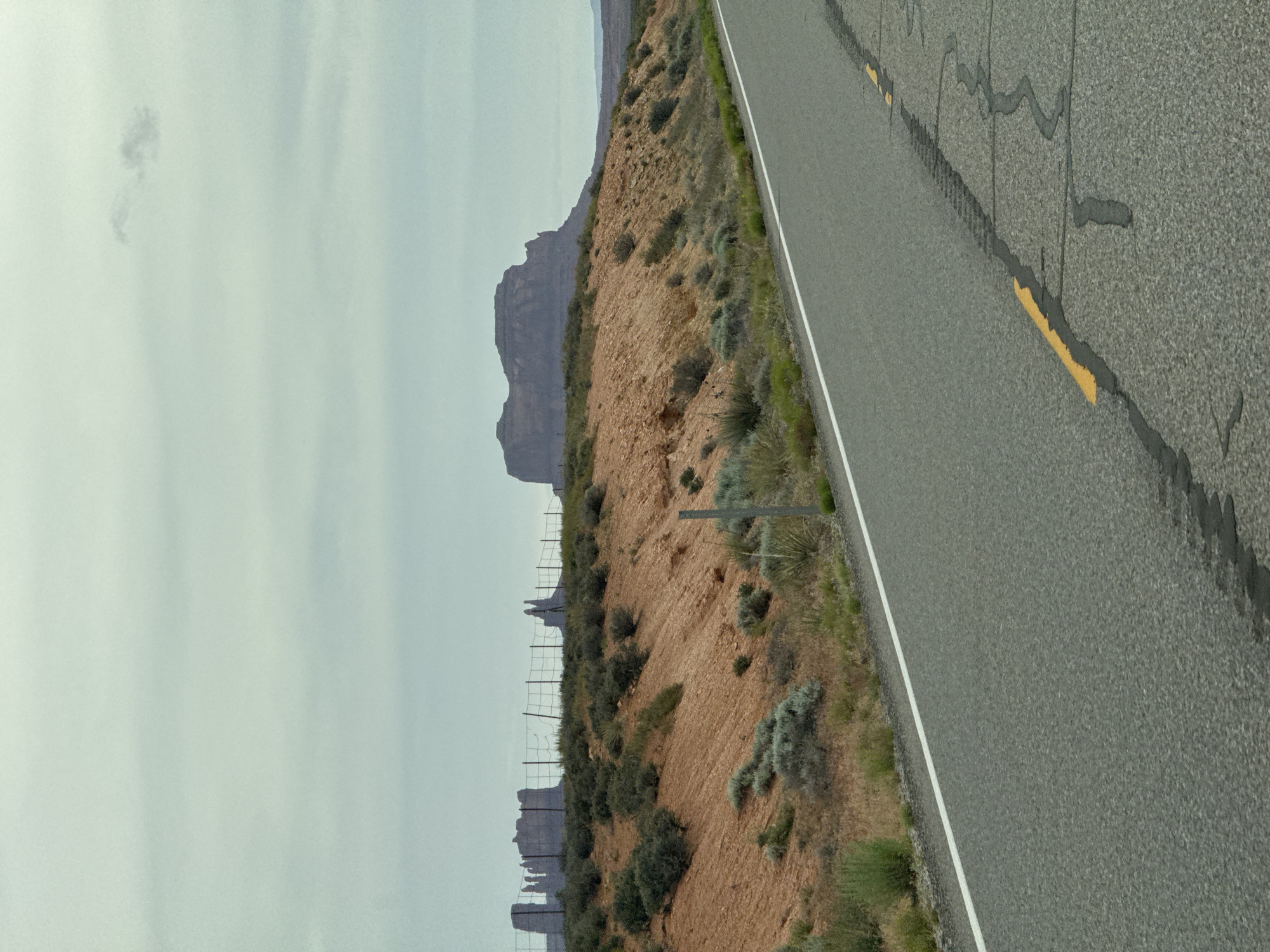 The image depicts a road running alongside a desert landscape. The road has visible lane markings and patches. On the left of the road, there is a reddish-brown hillside with sparse vegetation. In the distance, there are rock formations and a cloudy sky. The location is unknown.