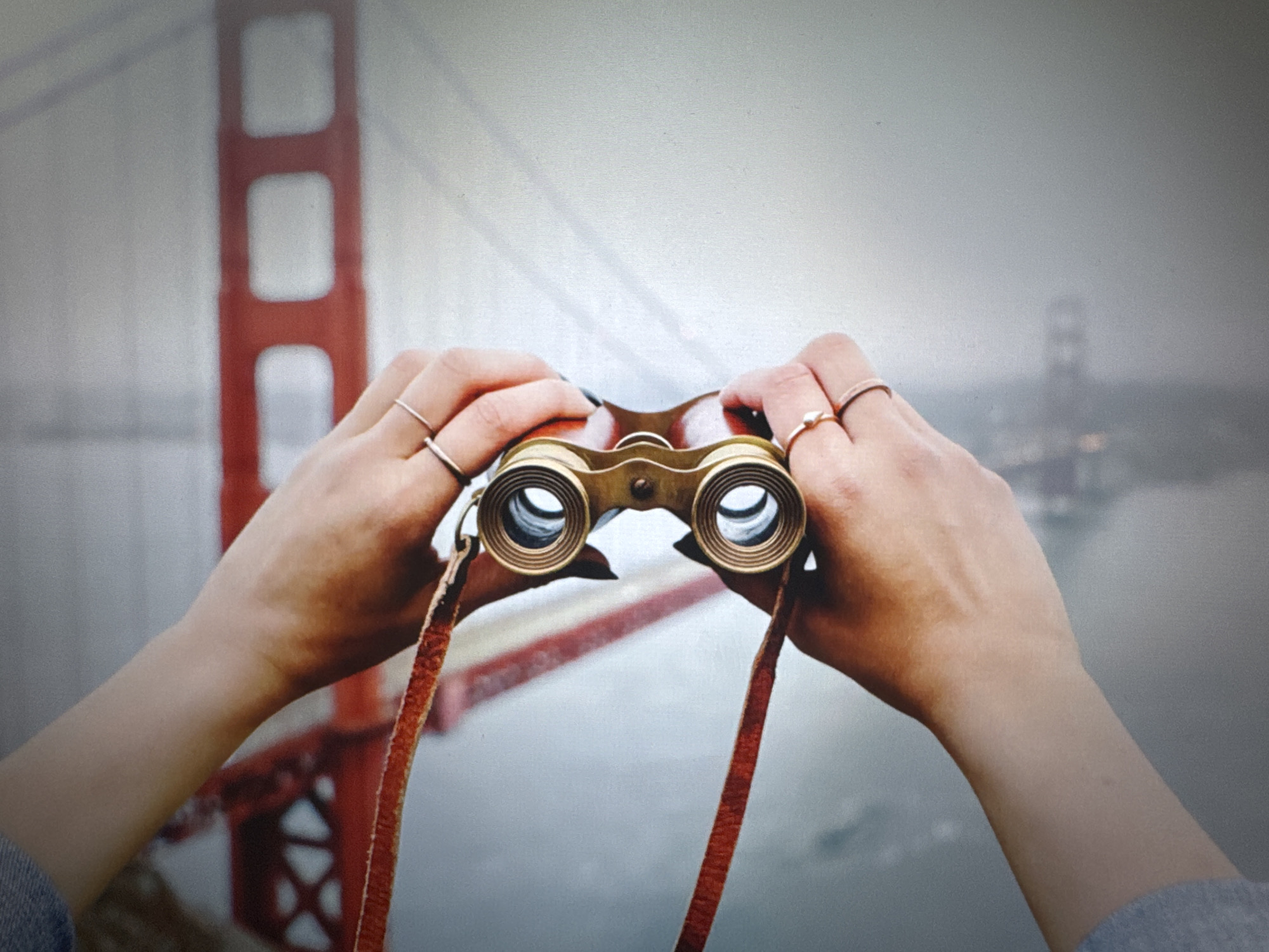 The image captures a first-person perspective looking over a pair of hands holding vintage binoculars. The hands, which appear to belong to a person wearing a light blue denim sleeve on the left arm and a grey sleeve on the right, are adorned with multiple simple rings on several fingers. The left hand has black painted fingernails, while the right hand has a light-colored, possibly diamond-shaped, ring on the ring finger.
The binoculars are made of brass or a gold-toned metal, featuring a classic design with a worn leather strap hanging downwards. The primary activity depicted is observation or sightseeing, as the binoculars are held in a viewing position.
In the background, the iconic red structure of the Golden Gate Bridge is visible, partially obscured by a thick layer of fog or haze. The soft, diffused lighting suggests an overcast day. The bridge's towers and cables are discernible, stretching across what appears to be a body of water, likely a bay. Another, more distant section of the bridge is faintly visible through the fog on the right side. The overall atmosphere is serene and somewhat mysterious due to the misty conditions. There is no visible text in the image.