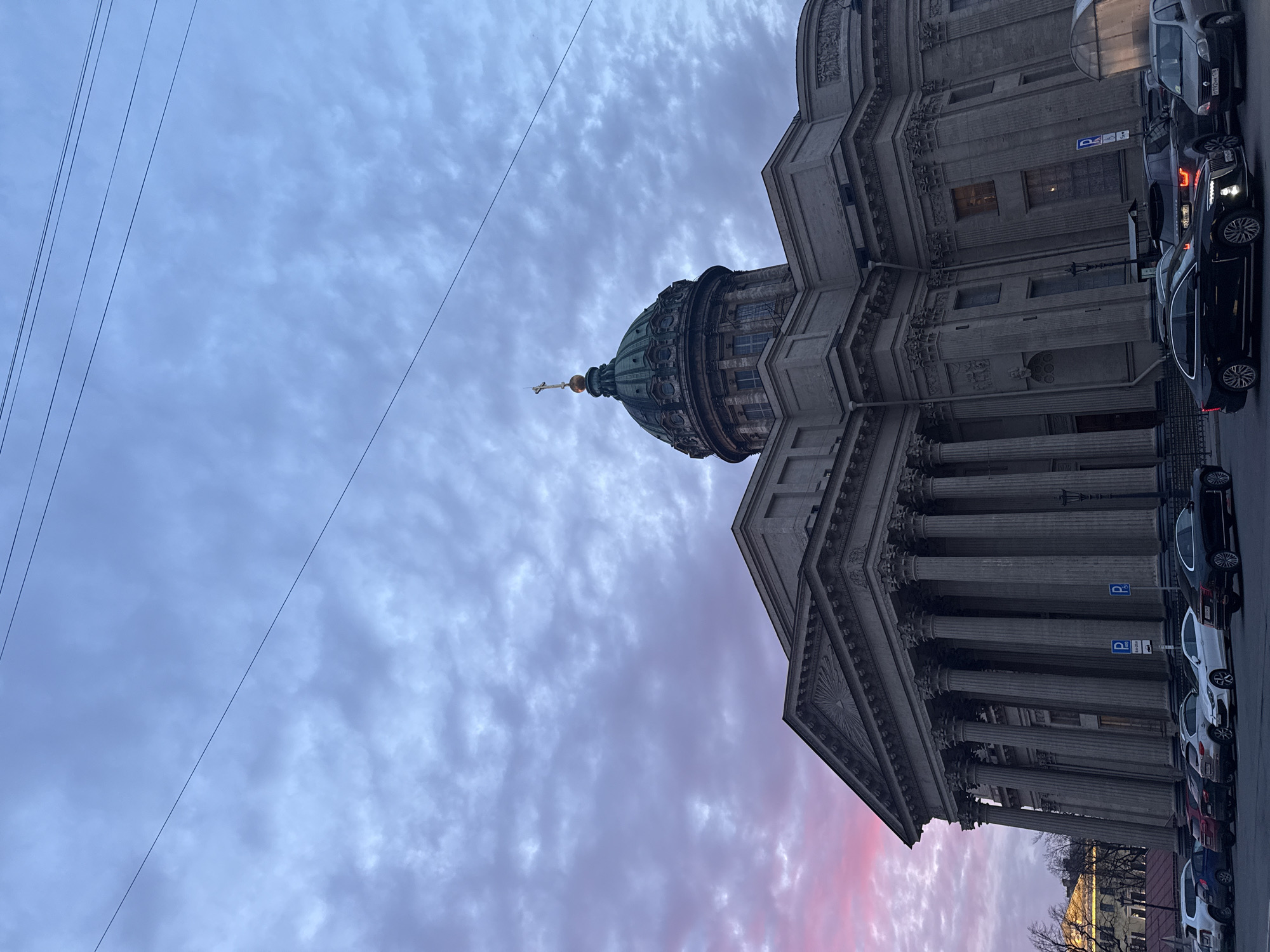 The image depicts the Kazan Cathedral in Saint Petersburg, Russia, from a low angle. The cathedral's dome and colonnade are visible against a cloudy, pink-tinged sky. Power lines stretch across the upper-left portion of the frame. Parked cars line the street adjacent to the building. No people are visible in the frame.