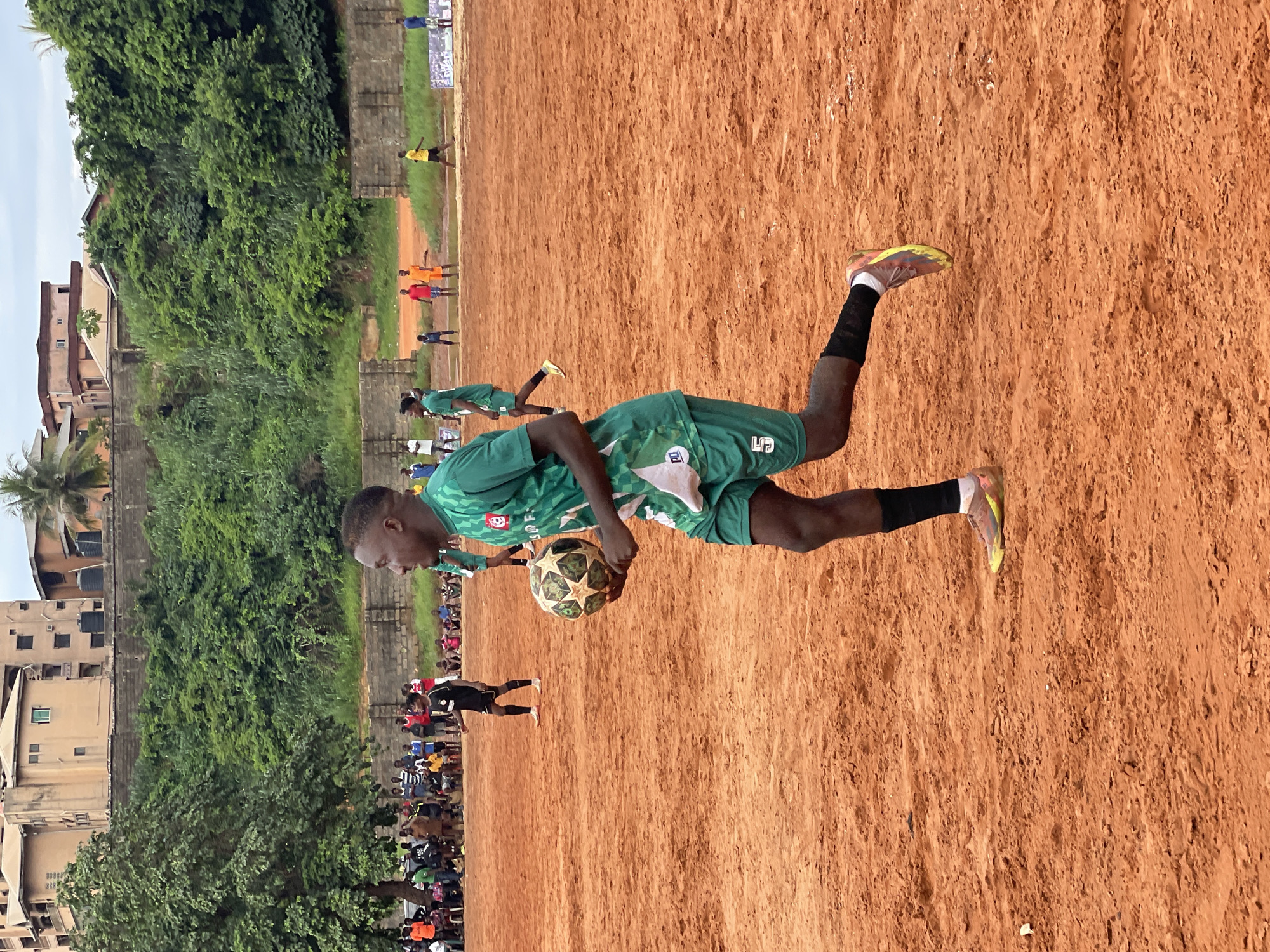 An outdoor, low-angle, medium shot captures a male soccer player in the foreground, holding a ball in his right hand. He is running to the right of the frame, on a dusty, reddish-orange soccer field. He is wearing a green soccer jersey with white accents and a white trim, green soccer shorts, black socks, and multi-colored soccer cleats. The ball has a star pattern and is predominantly white with brown accents. The player has dark skin and short black hair. In the background, several other players in various colored jerseys are visible, some sitting on the ground, and others standing. A line of spectators is seated on a bench behind them. Beyond the field, a dense green hillside covered in trees and shrubs rises up, with buildings visible at the top. A cloudy sky completes the scene.