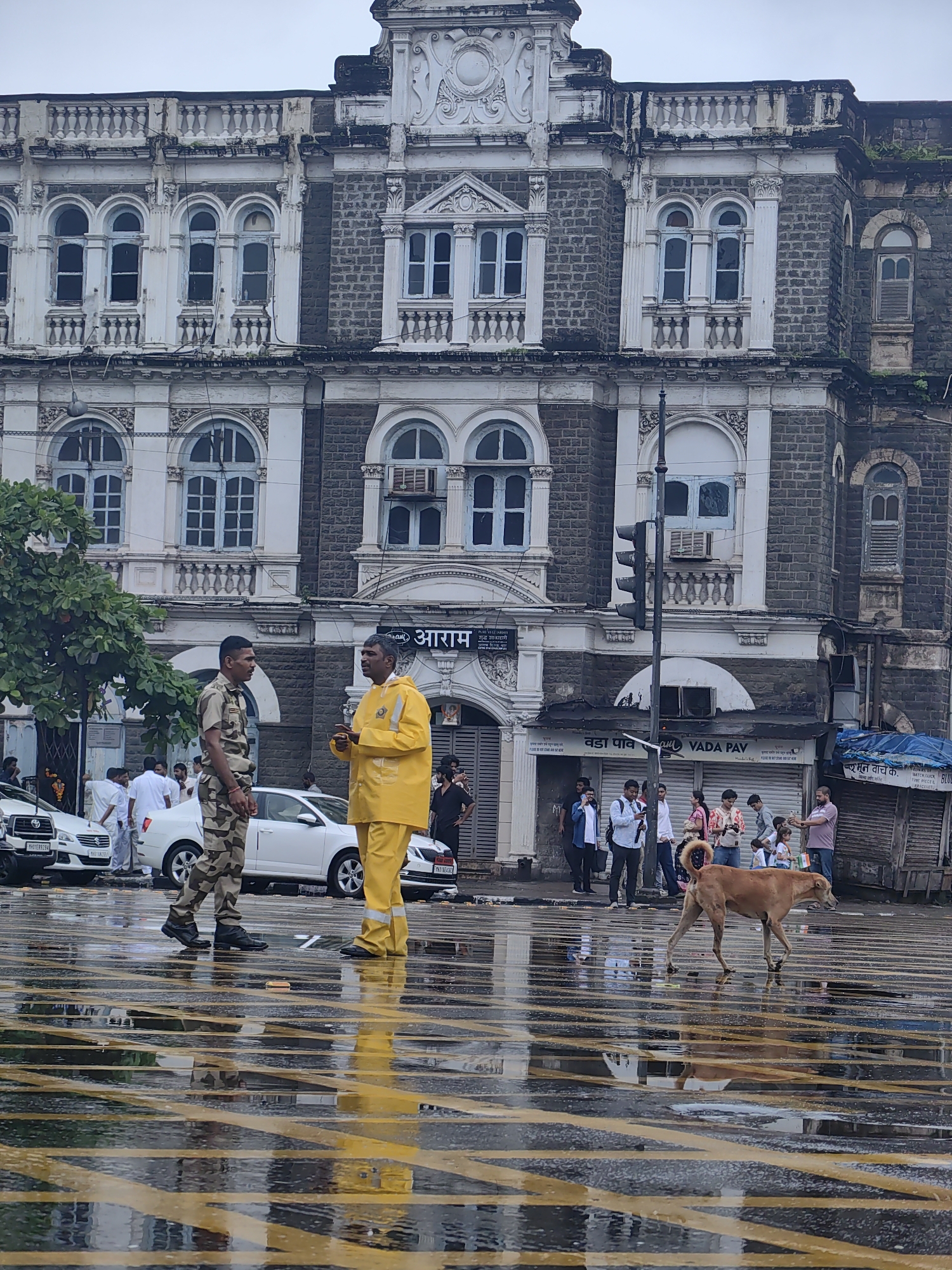 In Mumbai, India, on a rainy day, two men stand on a wet, yellow-lined street. One man, in a camouflage uniform, faces right. The other, wearing a bright yellow raincoat and pants, faces left, gesturing with his hands as he speaks. Behind them, a brown dog walks right to left. A large, ornate building with grey stone and white decorative elements dominates the background. Cars are parked on the street, and several people are visible in the distance, some under awnings. The street's surface reflects the overcast sky and surrounding structures, indicating recent rainfall. A traffic light stands to the right of the central figures, and various signs, including one for "Vada Pav," are visible on the storefronts.