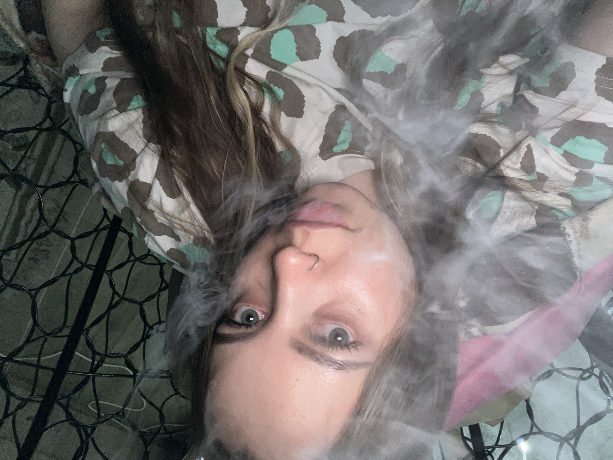 A person lies on their back with their face towards the camera, partially obscured by smoke. The person has fair skin, light-colored eyes, a nose piercing, and light brown hair. They are wearing a patterned shirt with light brown, dark gray, and green leopard spots. The person appears to be resting on a black metal wire frame or grid, potentially a chair or cot. A pink object is faintly visible in the lower right corner of the image, behind the smoke. The background is a drab, off-white color. There is no clear indication of the location of Novopokrovskaya, Russia.