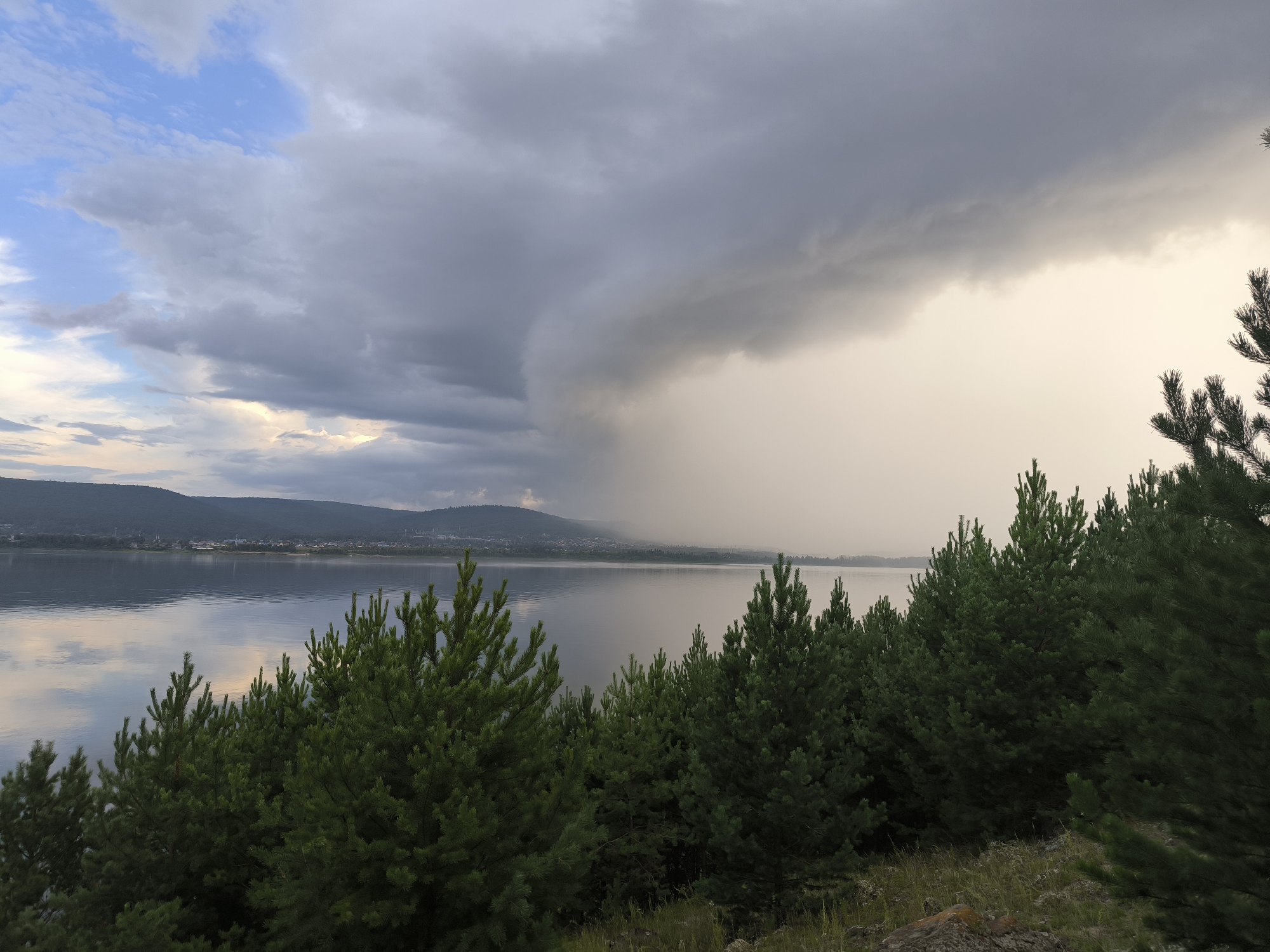 A wide shot captures a dramatic storm front approaching across a serene lake near the city of Greemuchiy, Russia. The foreground is dominated by a dense line of dark green pine trees, their branches silhouetted against the sky. Beyond the trees lies a calm, reflective lake, its surface mirroring the muted colors of the sky. In the distance, the low, rolling hills of the opposite shore are visible, dotted with buildings and structures of Greemuchiy. The sky is a turbulent mix of dark, heavy storm clouds on the left, transitioning to a lighter, hazy sky on the right, where a curtain of rain is visibly falling. The light is dim and diffused, creating a somber and awe-inspiring atmosphere.