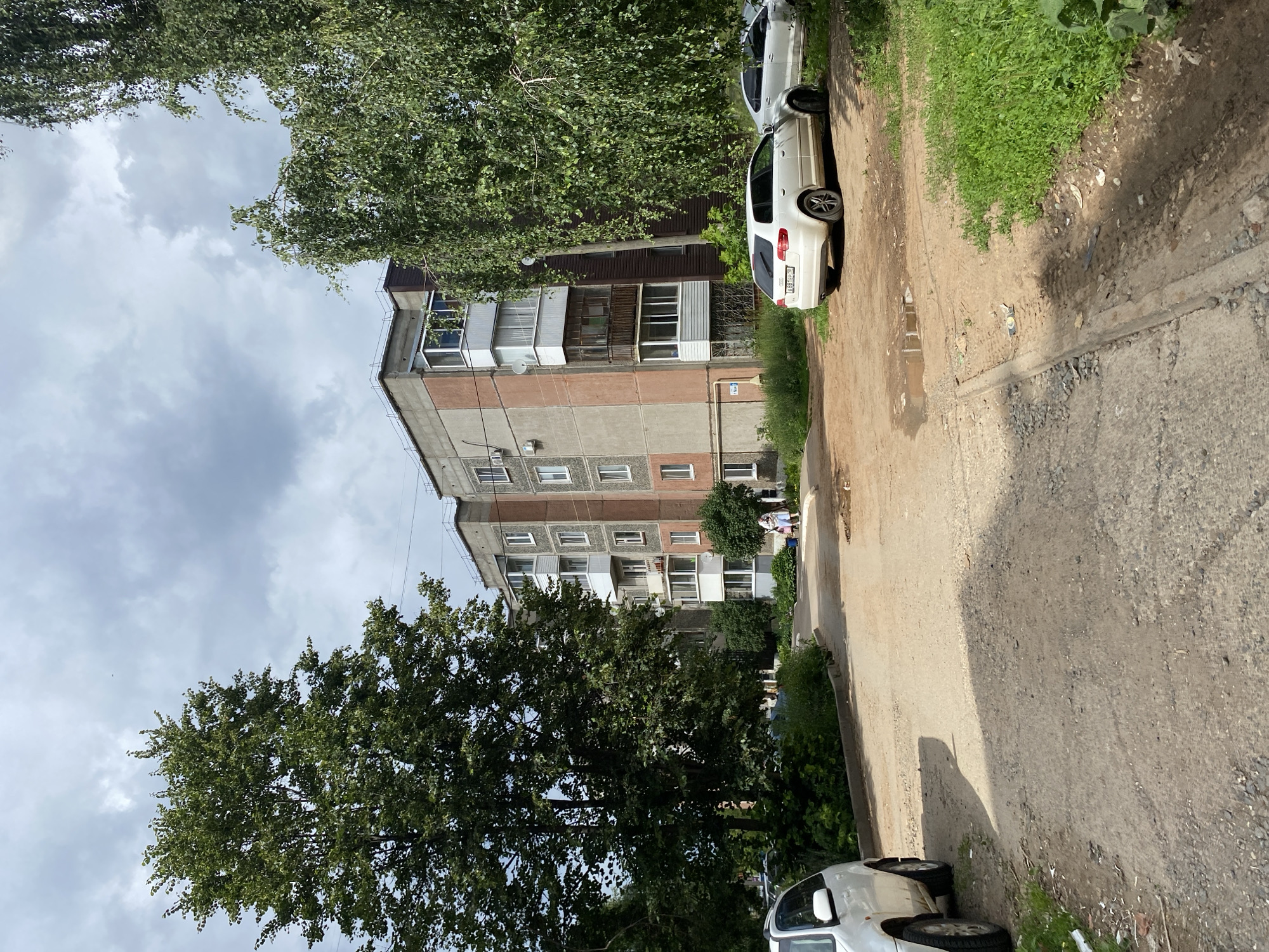 The image depicts a residential courtyard in Votkinsk, Russia, characterized by a multi-story apartment building and surrounding greenery. The building, appearing to be made of concrete and plaster, has a faded reddish-brown facade interspersed with grey sections and features numerous balconies, some enclosed with windows. Two white cars are parked on a dirt and gravel surface in front of the building. One car, a white hatchback, is closer to the foreground and is partially obscured by shadow. Another white car is parked further back. A dirt path with puddles of water is visible in the foreground, suggesting recent rainfall or a drainage issue. Lush green trees and bushes line the perimeter of the courtyard, with one large tree dominating the left side of the frame and another dense tree on the right. The sky is overcast with grey clouds. In the distance, near the building, two figures, possibly a woman and a child, are walking. The overall scene conveys a typical post-Soviet urban residential environment.