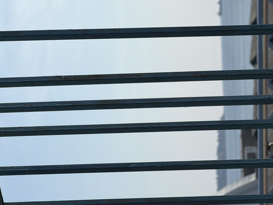 The image depicts a scene viewed through five vertically oriented, dark teal metal bars, exhibiting minor rust and wear. The bars dominate the immediate foreground, with the background significantly out of focus. The upper portion of the background reveals a clear, light blue sky. Below the sky, a body of water, likely a sea or large bay, extends to a distant horizon. On the far left of the horizon, a faint landmass with indistinct vegetation or structures is visible. Two blurry vessel silhouettes are present on the water in the mid-distance. Directly behind the vertical bars and below the water line, a segment of a light-colored structure appears on the left, and an outdoor air conditioning unit is discernible on a rooftop surface. A horizontal, light-colored railing traverses the lower section of the frame, positioned behind the vertical bars. The location is in the city of Unknown, Unknown.
