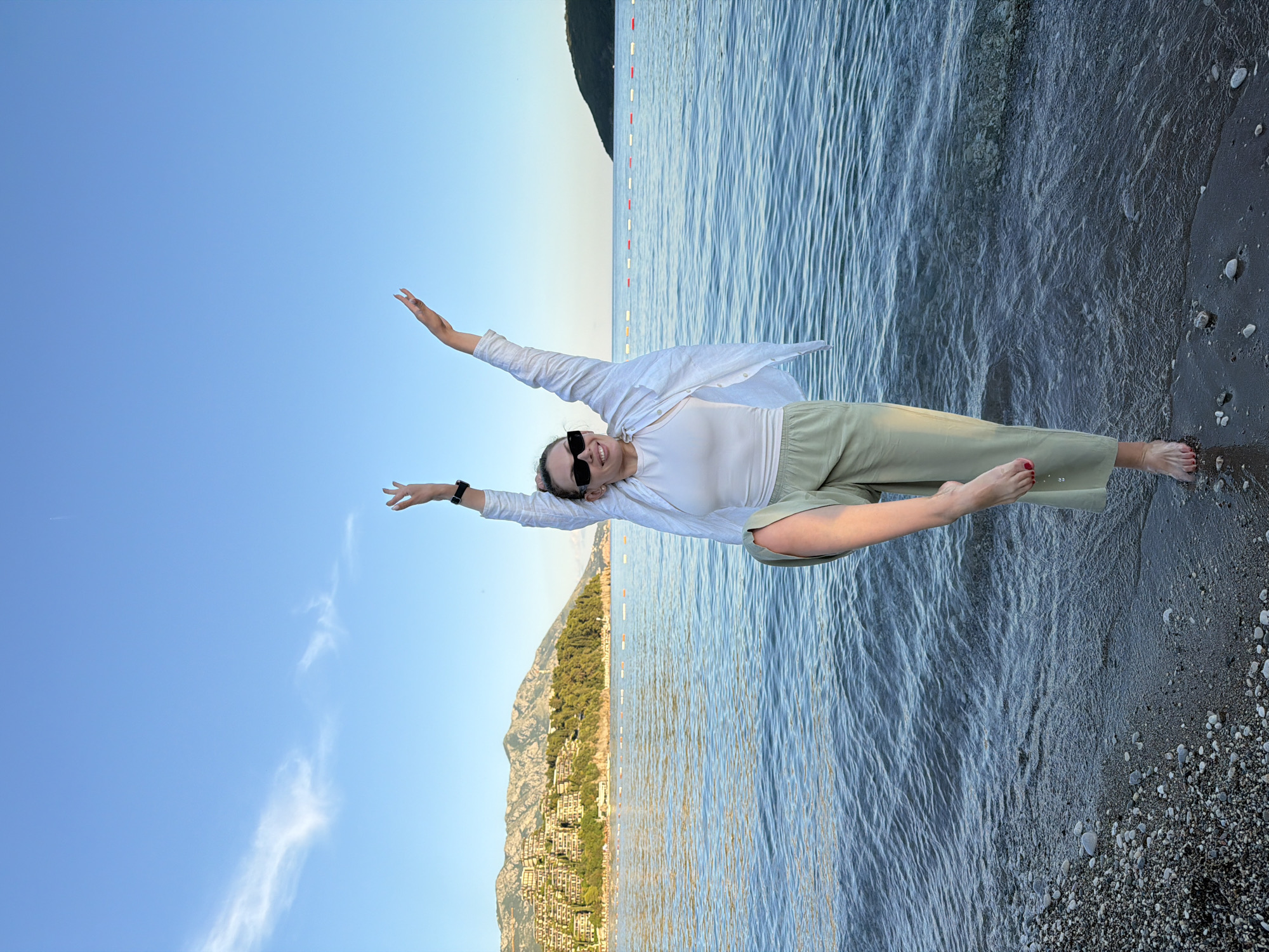 A young woman stands on a pebble beach in Budva, Montenegro, with the Adriatic Sea and a mountainous coastline in the background. She is wearing a white tank top, a white unbuttoned shirt, and light green wide-leg pants. Her left leg is bent at the knee, and her arms are raised overhead. The sea is calm, with gentle waves lapping at the shore. The sky is a clear blue with a few wispy clouds. The coastline is lined with trees and buildings, and mountains rise in the distance. The image is taken from a low angle, looking up at the woman and the sky.