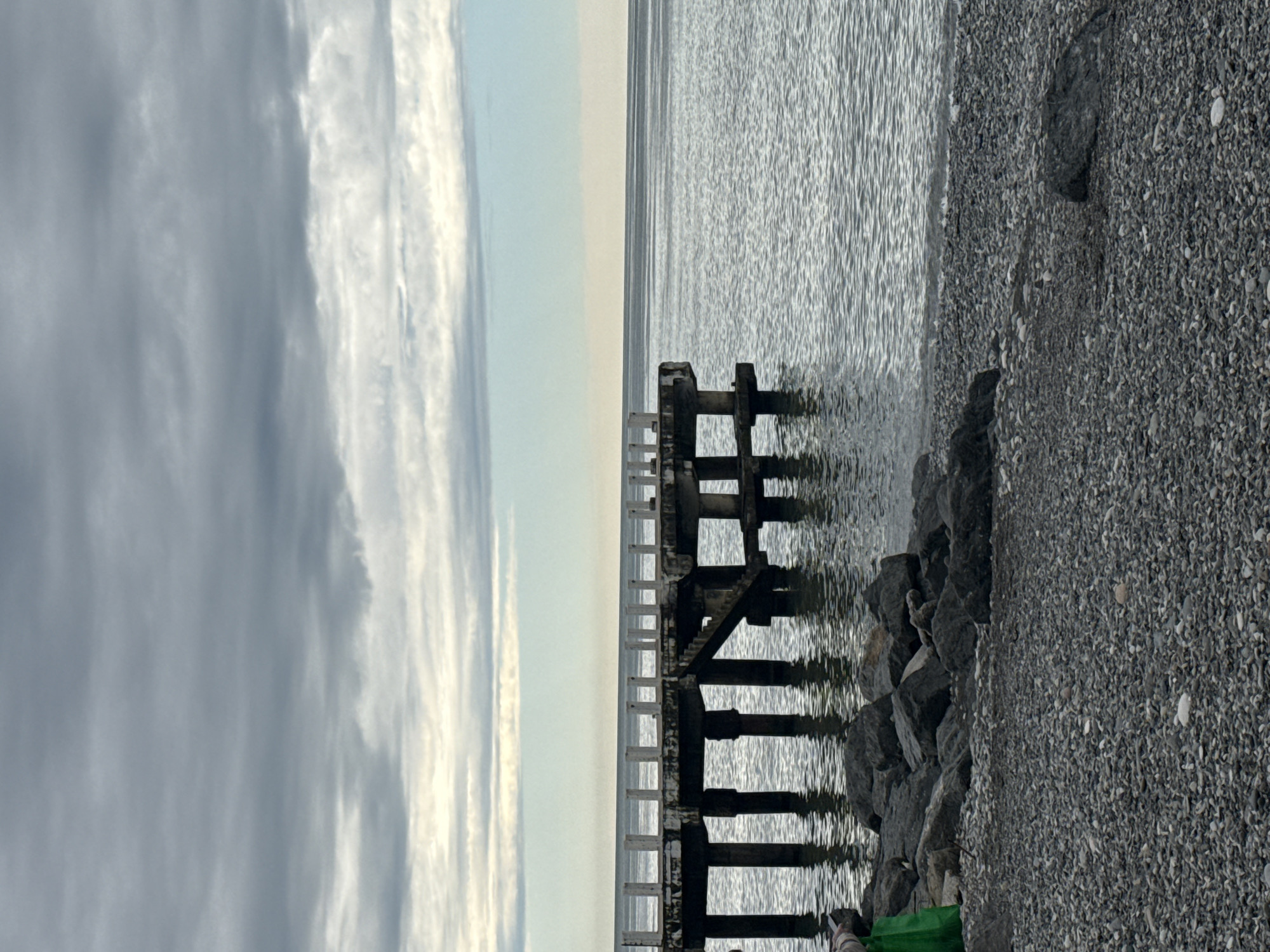 A vertical photograph captures a desolate coastal scene, likely in Batumi, Georgia, during overcast weather.
Foreground: A stony beach, composed of small, grey pebbles, occupies the right side. Larger dark rocks are positioned closer to the waterline. A portion of an object is visible at the bottom center of the frame.
Middle Ground: An old, partially submerged pier runs from the beach toward the horizon. The pier's decaying structure is evident, with visible posts and crossbeams.
Background: The grey water of the Black Sea stretches out toward the horizon, meeting the sky. The horizon line is a narrow band of pale light. The sky is filled with dense, grey clouds, with slight variations in shading and texture.
Overall: The image conveys a sense of solitude and decay, with the weathered pier and overcast sky contributing to a somber atmosphere.