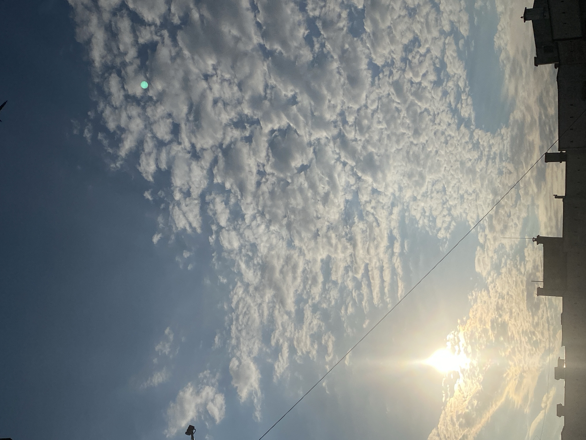 The image shows a sky filled with scattered cumulus clouds. The sun is setting on the left side of the frame, casting a warm glow and illuminating the clouds. In the foreground, the silhouettes of rooftops and building structures are visible, suggesting an urban environment. A thin wire extends diagonally across the sky. The overall scene is a view of the sky over the city of Pleven, Bulgaria, likely captured during the late afternoon or early evening.