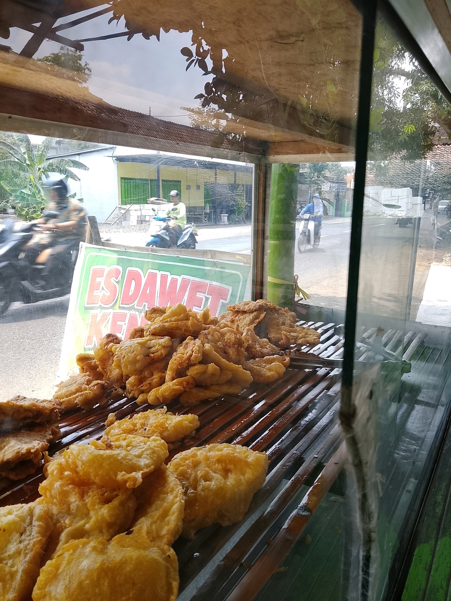 A street-side food stall in Banaran, Indonesia, is depicted through a glass display. In the foreground, a variety of fried snacks, appearing to be banana fritters and tempeh, are arranged on a wooden grate. A sign with red and white lettering reads "ES DAWAET KEM". Behind the food display, the street is visible with traffic, including two individuals on motorcycles. One motorcycle has a rider in a green shirt, and another has a rider in a blue jacket. Buildings with green doors and tiled roofs line the street. Trees and foliage are present in the background, indicating an outdoor setting. The overall lighting suggests it is daytime.