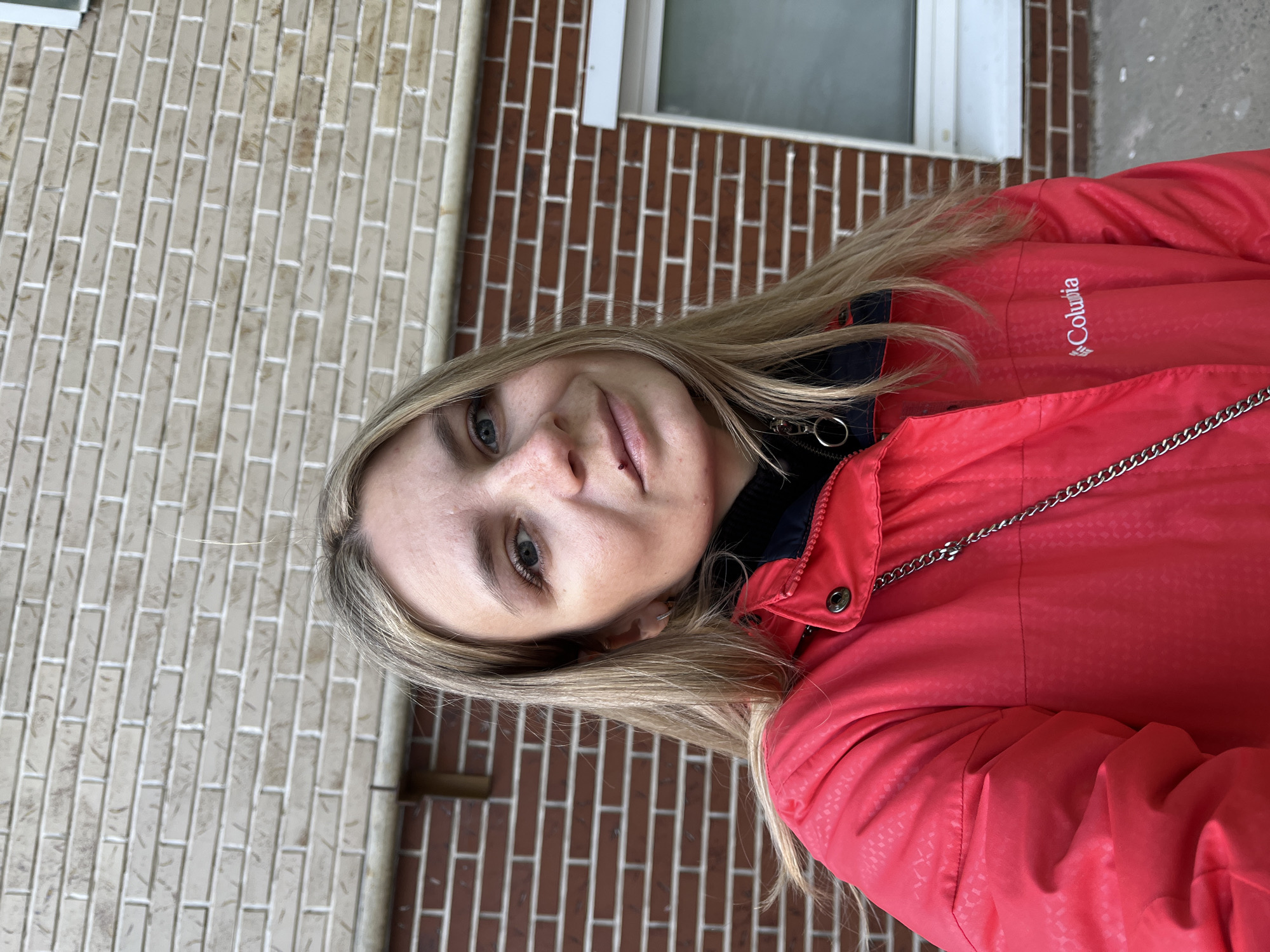 A blonde-haired woman wearing a red Columbia jacket is standing in front of a brick wall and a window. The location is Chaikovsky, Russia.