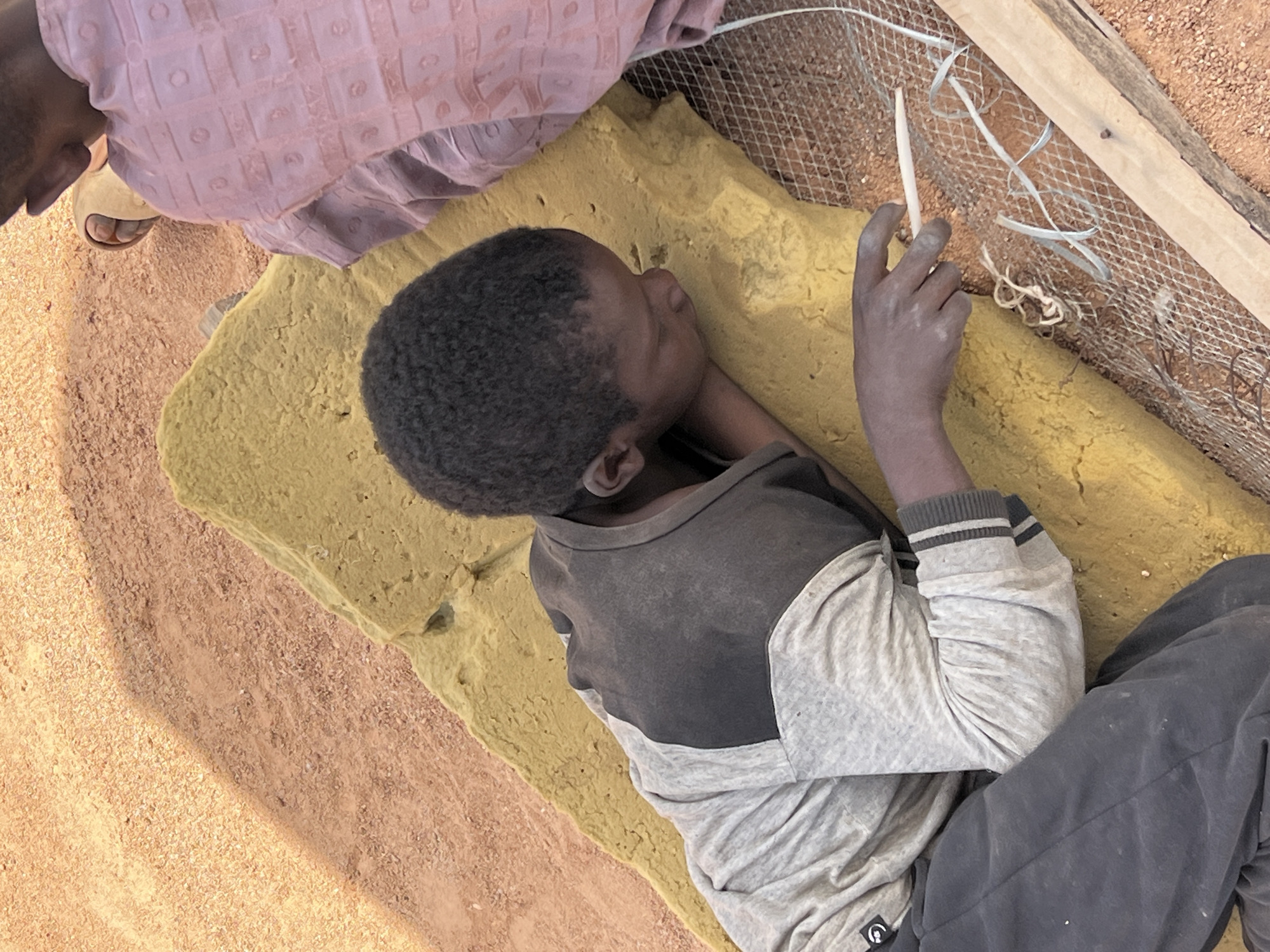 A person with dark skin and short, dark hair is lying on a yellow cushion. They are wearing a gray and black long-sleeved shirt and dark gray pants. The person is holding a white object in their right hand. The background includes a pink garment worn by someone else, sandy ground, and a wooden frame with wire mesh. The location is the city of Barakin Lerawi, Nigeria.
