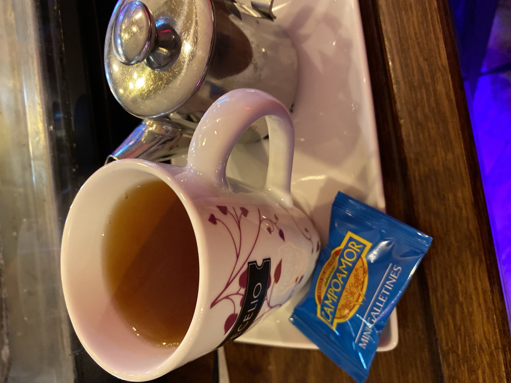 A cup of tea rests next to a small, blue packet of Campoamor Mini Galletines (biscuits) on a white square dish. The cup is predominantly white with a decorative burgundy pattern featuring stylized vines and a ticket-like shape bearing the word "CELIO". A silver teapot stands partly visible behind the cup. The setting appears to be a table, possibly in a restaurant or café, with a portion of a dark wooden surface visible. Given the mention of Campoamor Mini Galletines, it is likely in Spain, and the prompt indicates it's in Gijón/Xixón.