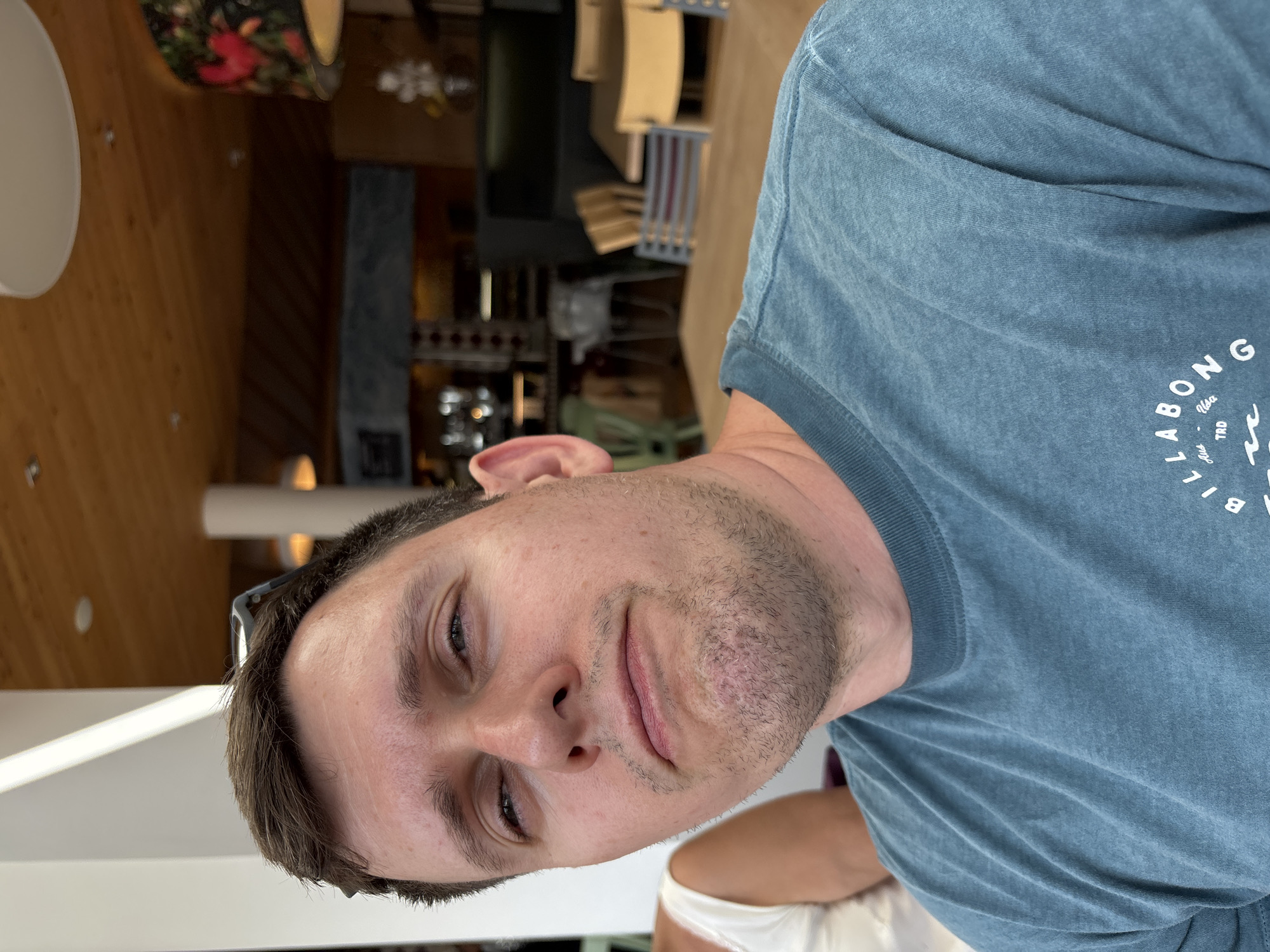 The image shows a medium shot of a man with fair skin and brown hair, looking upwards and to his left with a neutral expression. He is wearing a blue t-shirt with a logo that reads "BILLABONG TRD Aus Usa". He appears to be sitting at a wooden table in an indoor setting with a warm-toned wooden ceiling and a chandelier with a floral pattern. The background suggests a restaurant or cafe with other tables and chairs visible. The man's left shoulder and arm are visible, as is a portion of another person's shoulder wearing a white top. The scene does not provide any specific information about the city of Heiligenblut am Großglockner, Austria.