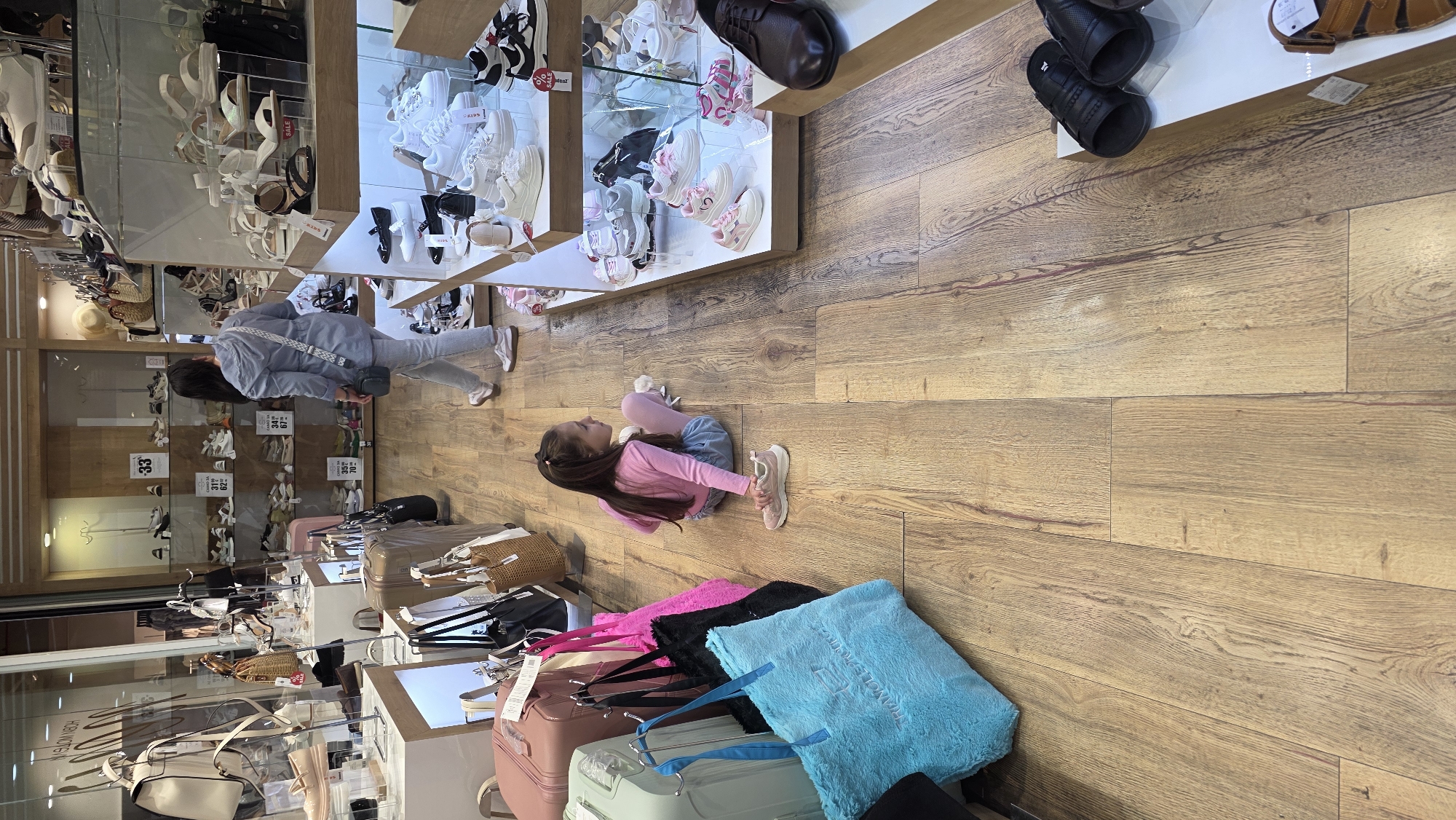 A medium-angle, indoor shot shows a shoe and luggage store. The scene has a little girl and an adult looking at shoes in a display cabinet.
The little girl is sitting on the wooden floor and is wearing a pink long-sleeve shirt, blue jeans, and pink shoes. The adult is wearing a grey long-sleeve shirt and blue pants and is standing, looking at the display cabinet with shoes.
There are two shoe display cabinets in the store. The upper cabinet has shoes on display, while the cabinet below has more shoes and price tags. In the bottom left corner of the image is a luggage display with multiple colored pieces of luggage and bags.
The image has a neutral color palette and is well-lit. The scene appears to be taken in a retail store in Blagoevgrad, Bulgaria.