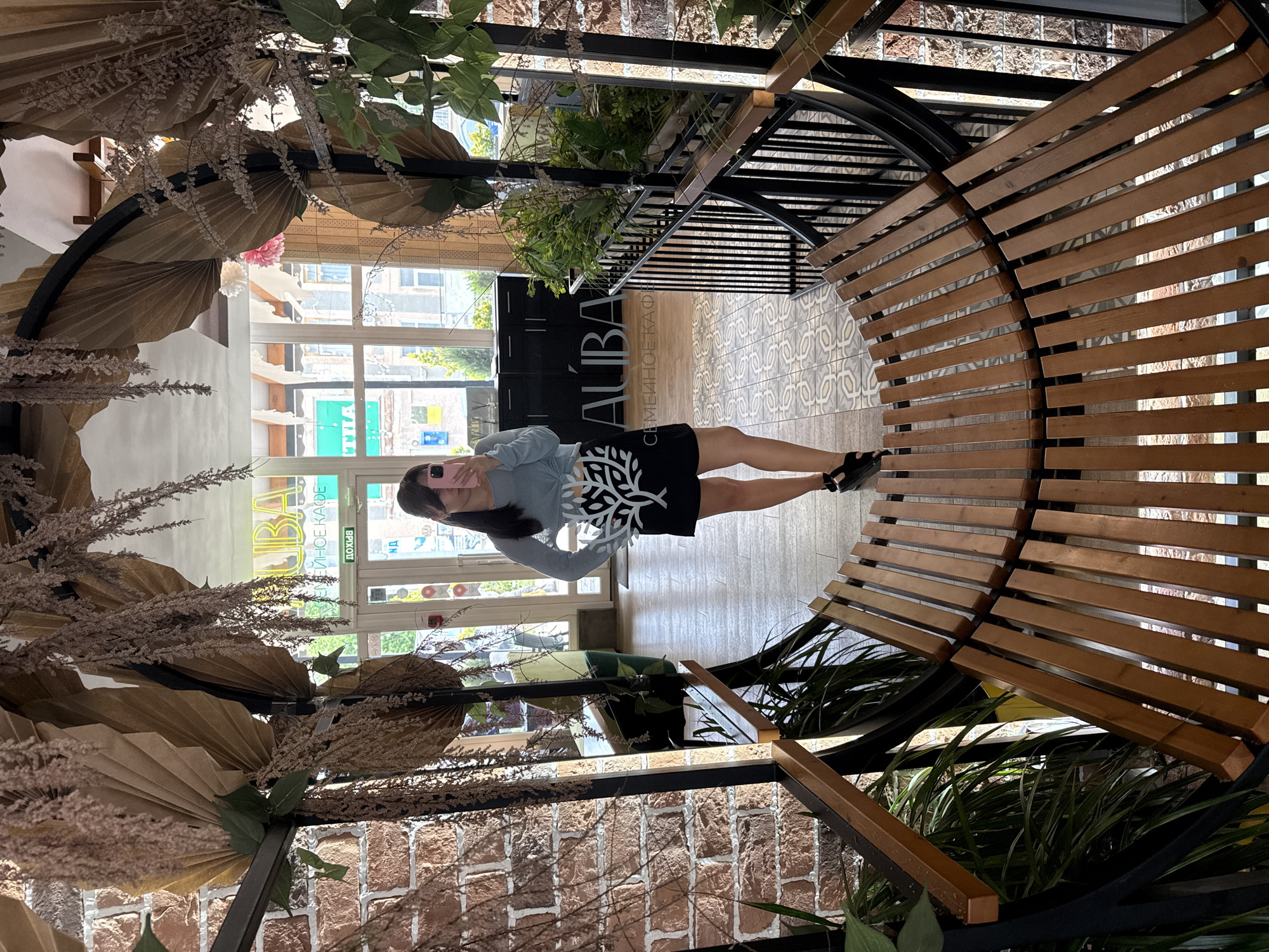 A woman stands on a wooden bench in front of a mirror, taking a selfie. She is wearing a light blue long-sleeved shirt and a black skirt with a white tree pattern. The bench is curved and made of wooden slats. Behind her, a brick wall is visible, and in the mirror, an interior of a cafe is reflected. The cafe has a sign that reads "AUBA" and "СЕМЕЙНОЕ КАФЕ" (Family Cafe). The reflection also shows windows and plants. Dried decorative branches and leaves hang from the ceiling. The floor has a patterned tile design.