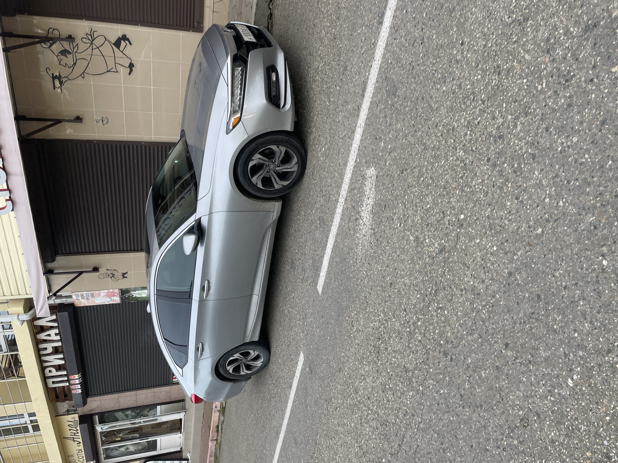A silver Honda Accord sedan is parked in a parking space adjacent to a building in Olginka, Russia. The building has a brown metal roll-up door with a sign that includes the word "ПРИЧАЛ" in Cyrillic. There is artwork on the building wall above the door depicting a chef. The parking space is marked with white lines on an asphalt surface.