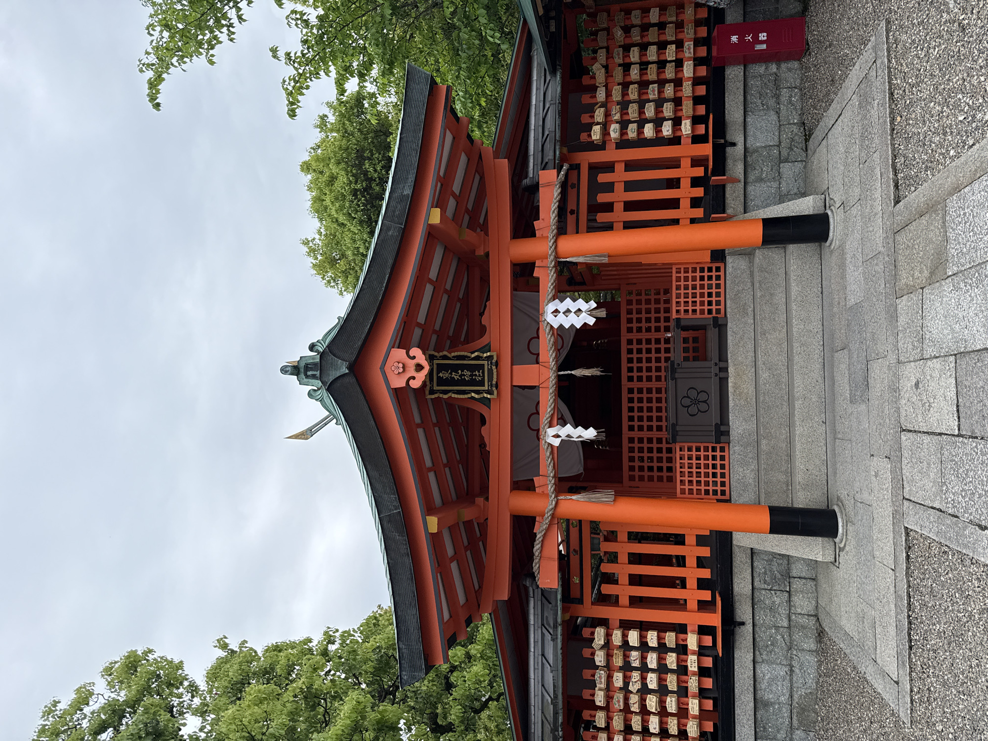 A vertical, low-angle photograph shows a Japanese Shinto shrine, likely located in Kyoto. The shrine is predominantly red, featuring a traditional sloping roof with dark gray or black trim. A sign with Japanese characters hangs above the entrance. The shrine is decorated with white paper zigzags. Wooden ema (votive tablets) are attached to the side walls of the shrine.
The shrine is set on a stone foundation, with a stone staircase leading to the entrance. Green trees are visible in the background. The sky is overcast. A red fire extinguisher is affixed to the side. The image provides no evidence of human presence.