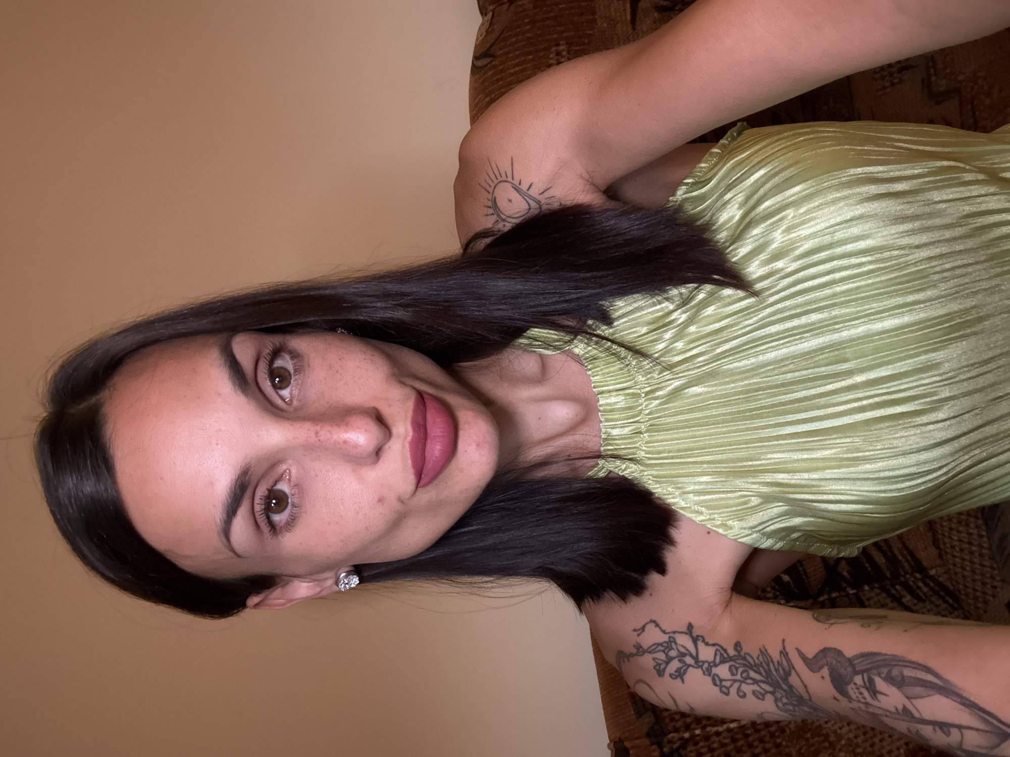 A medium shot shows a woman with dark hair and brown eyes, wearing a light green halter top and a diamond stud earring. She has several tattoos on her arms, including a detailed drawing of a woman with horns on her left arm and a sun-like symbol on her right shoulder. Her lips are painted a matte pink color. The background is a plain, light brown wall. The woman is seated on a patterned couch. The scene is indoors and appears to be a residential setting. There is no information in the image to suggest a location in Pleven, Bulgaria.