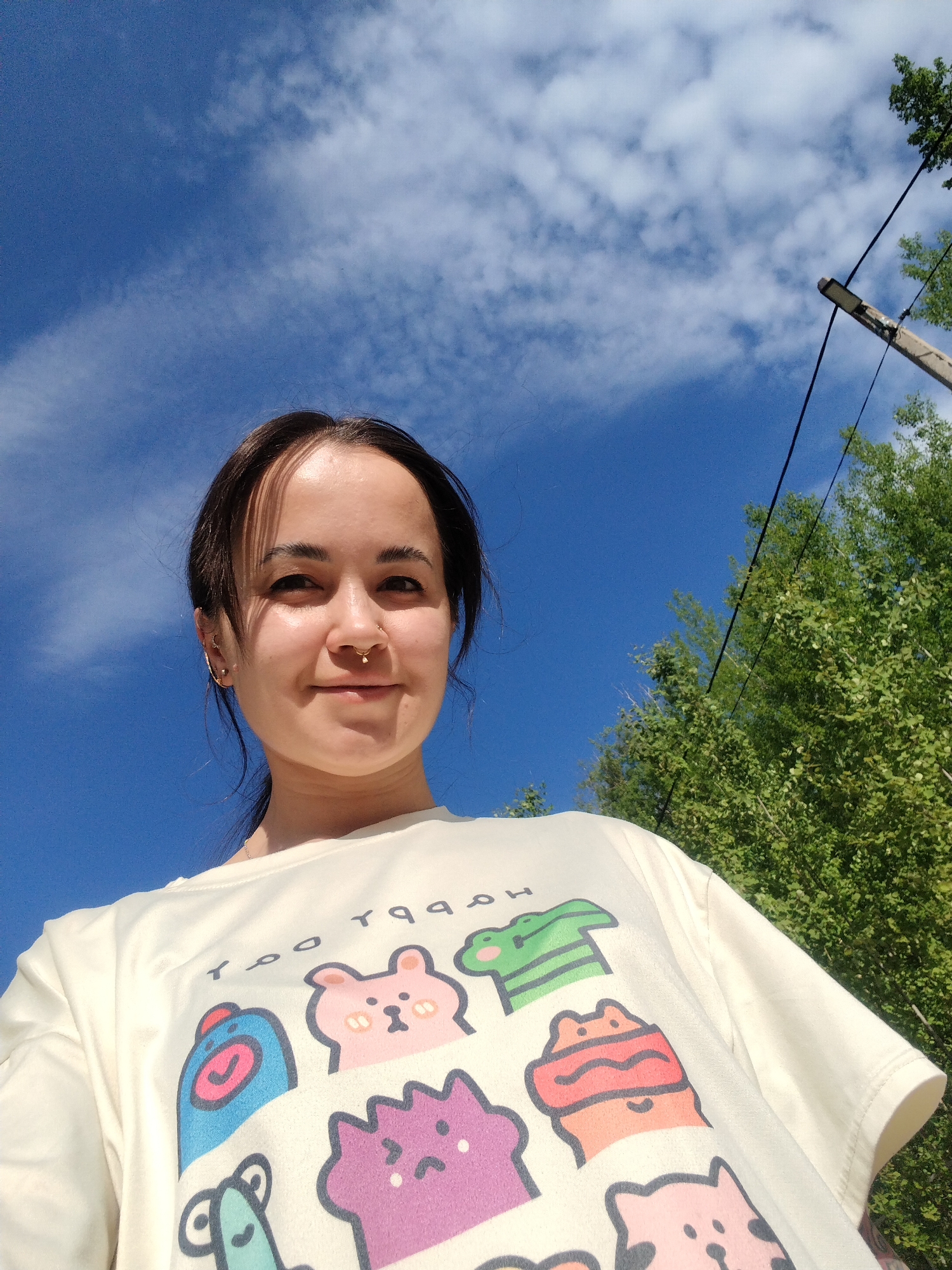 A young woman stands outside on a sunny day, looking directly at the camera. She wears a light-colored t-shirt with cartoon animal illustrations. The background features a bright blue sky with scattered clouds, a wooden utility pole with wires, and green foliage. The location is Noviy, Russia.