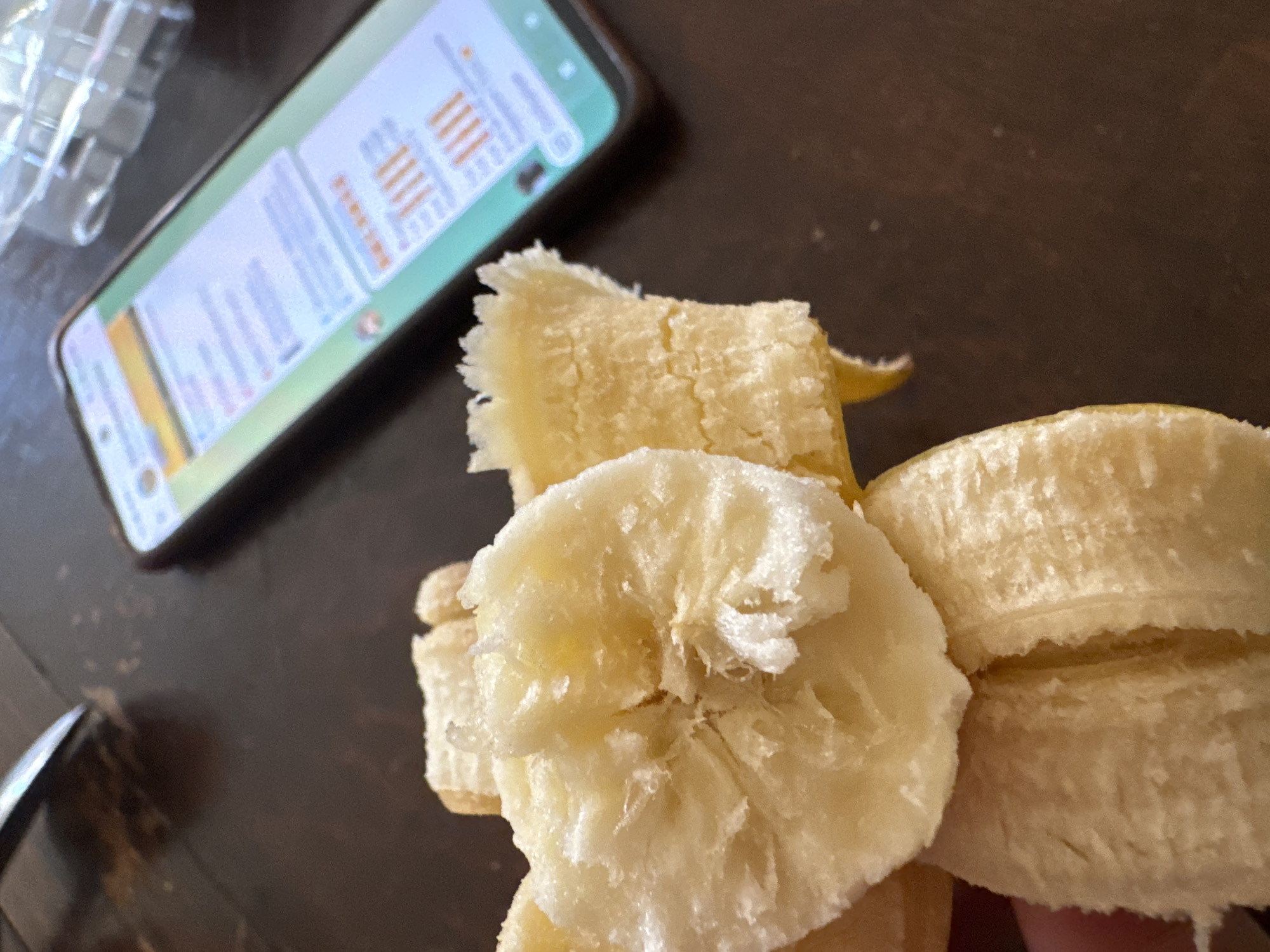 A close-up shot features a peeled banana, broken into several pieces, with the fragmented pulp prominently displayed in the foreground. In the background, a smartphone with an illuminated screen rests on a dark, wooden surface. The screen displays what appears to be a social media feed or a messaging application, with text and graphics visible. The overall scene suggests a casual moment, possibly at a table. No specific location in Waterford, Ireland, can be identified from the provided image.