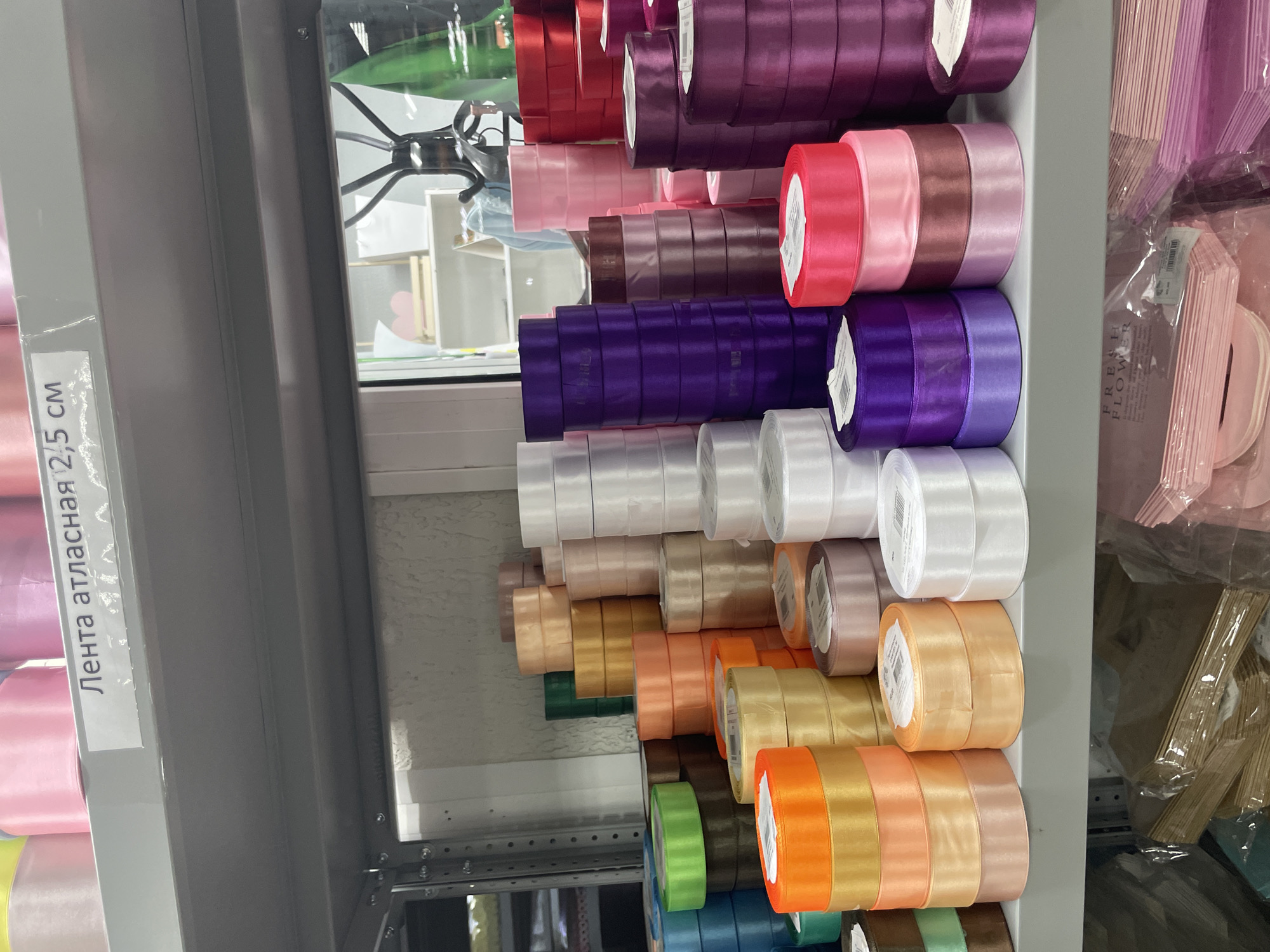 The image shows shelves stocked with rolls of satin ribbon in a variety of colors. The ribbons are neatly stacked by color, ranging from shades of red, pink, purple, white, tan, orange, green, and blue. The shelves are gray and appear to be part of a retail display, with the ribbons arranged for sale. A white label with Cyrillic text "Лента атласная 2,5 см" is visible on the shelf. Other packaging is visible to the right of the ribbons. The location is Unknown, Unknown.