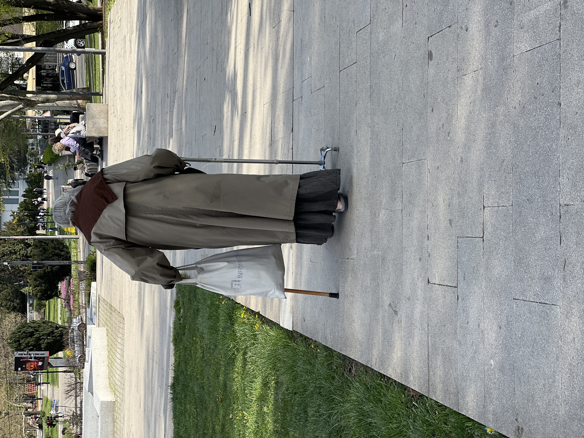 A street-level, medium-shot captures an elderly person from behind, strolling along a paved path in what appears to be a park or public space in the Palilula Urban Municipality, Serbia. The individual is dressed in a long, light olive-green coat, with a dark brown scarf or collar, and a dark pleated skirt or dress showing beneath the coat. They have gray hair and are using a silver-colored walking stick. They are also carrying a white cloth bag in their left hand.
The path is made of large gray stone tiles. To the side of the path is a grassy area. In the background, there are other people, trees, buildings, and parked cars, suggesting an urban environment. The lighting indicates it is daytime, with shadows cast on the path. A billboard is visible in the distant background with a colorful advertisement.
