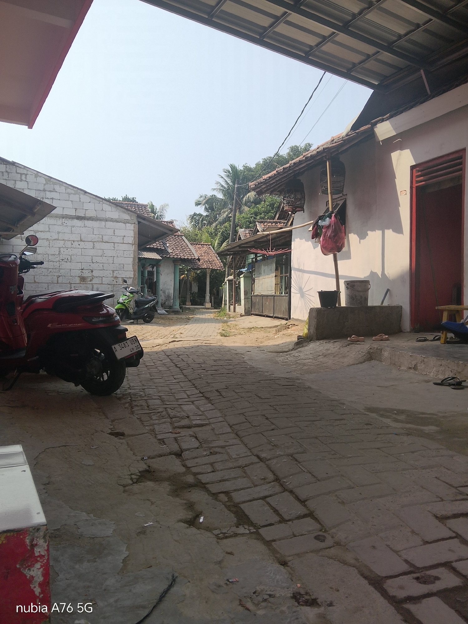 The image depicts a street scene in Cakung, Indonesia, characterized by residential buildings and paved pathways. A red scooter is parked in the foreground on the left, with its rear visible. Further down the street, a green scooter is parked. The buildings are predominantly single-story structures with tiled roofs, some exhibiting signs of wear and tear. The pathway is paved with bricks, and there are some puddles of water on the ground, suggesting recent rain or a damp environment. Vegetation, including palm trees, is visible in the background, indicating a tropical climate. Shadows are cast on the ground and buildings, suggesting it is daytime with sunlight. There are no people visibly present in the image. The overall atmosphere is one of a quiet, everyday street in a residential area.