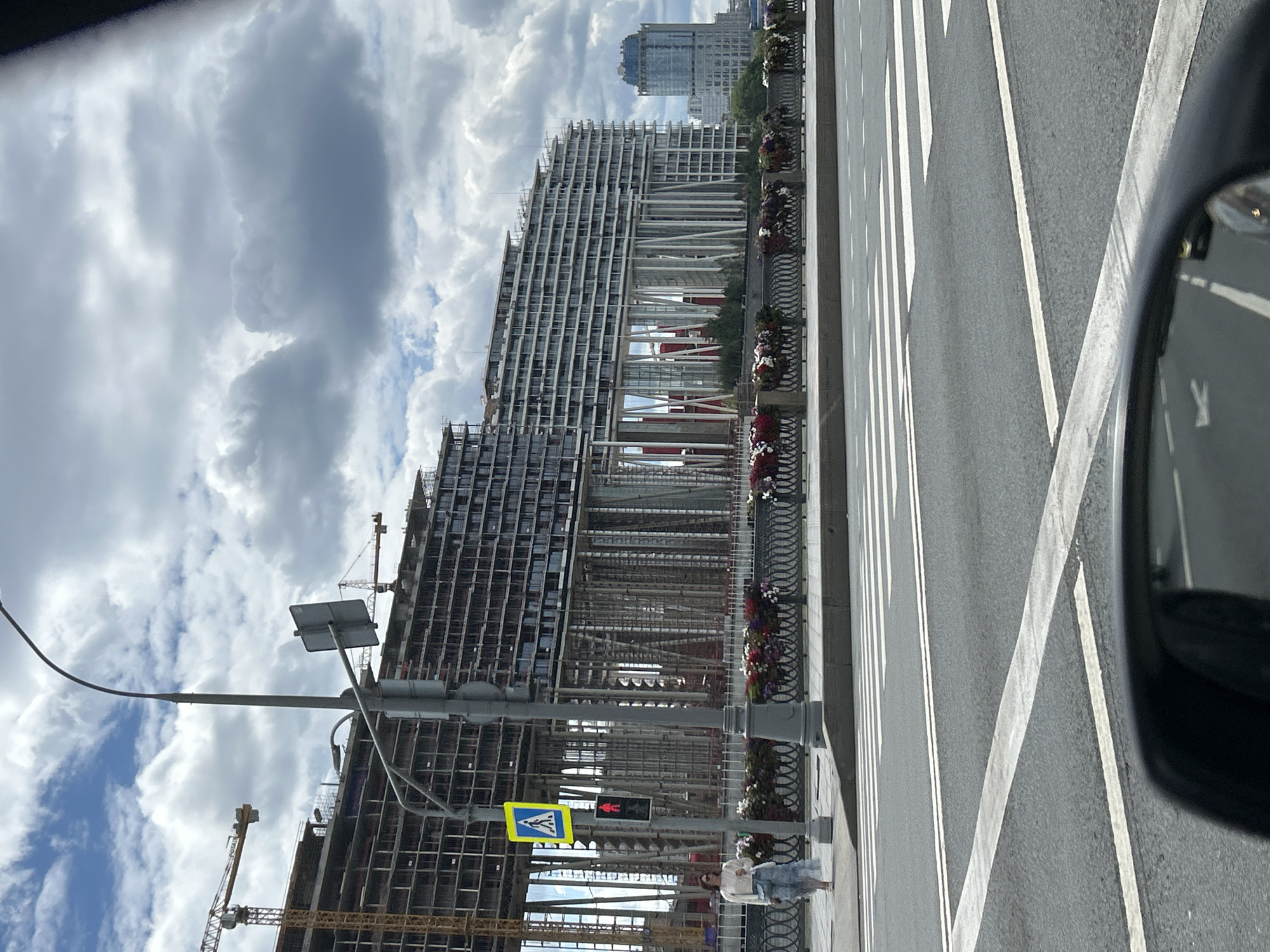 The image depicts a street scene in Moscow, Russia, featuring a massive, partially constructed building that dominates the background. The building appears to be a multi-story residential complex, characterized by its repetitive facade of windows and a complex structural framework with numerous supporting columns. Several construction cranes are visible around and above the building, indicating ongoing work.
In the foreground, a woman is walking on a sidewalk next to a street. She is wearing a light-colored top and a long skirt, and carrying a bag. A traffic light with a pedestrian crossing signal is located on the sidewalk, and a pedestrian crossing is marked on the road. The street is paved, and white lines delineate lanes and the crosswalk.
The environment includes a cloudy sky with patches of blue. A decorative metal fence with planters filled with flowers runs along the edge of the sidewalk, separating it from the road. In the distance, other buildings, including a taller, modern-looking structure with a rounded blue-tinted top, are visible. The image seems to be taken from inside a vehicle, as a portion of a rearview mirror is visible at the bottom of the frame. The overall impression is one of urban development and construction in a metropolitan area.
