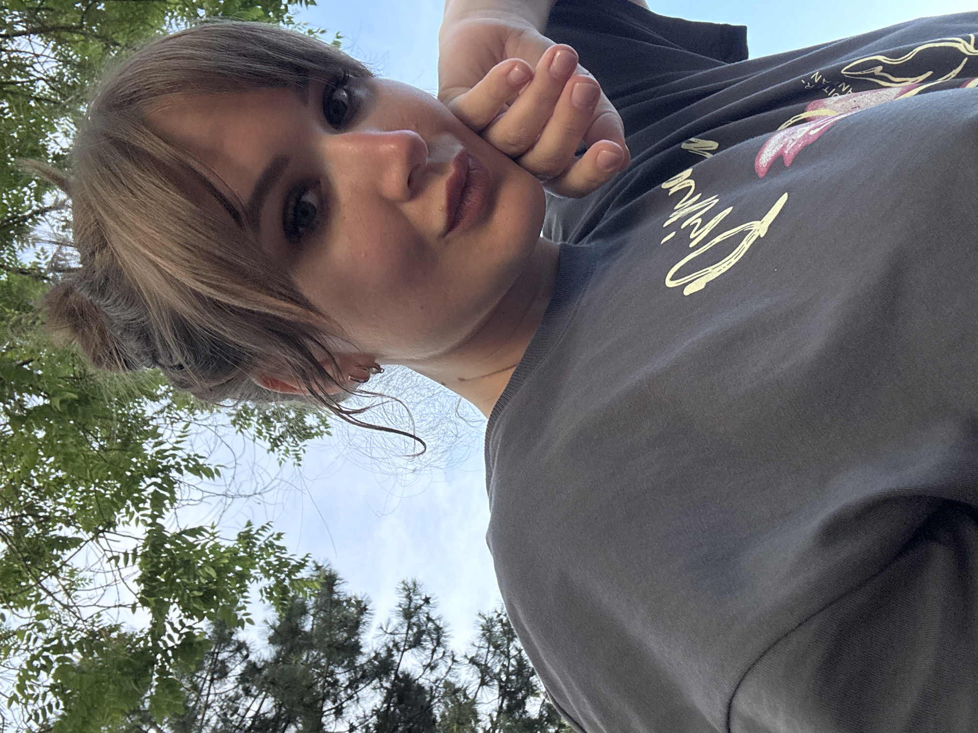 A young woman with fair skin, brown eyes, and light brown hair pulled back in a bun is seen in a low-angle, close-up selfie. Her right hand is placed against her cheek. She is wearing a gray t-shirt with a graphic design of white lettering and pink and yellow elements. The background includes green trees and a light blue sky with some white clouds. The specific location within Yevpatoriya, Ukraine, cannot be determined from the visual information provided.