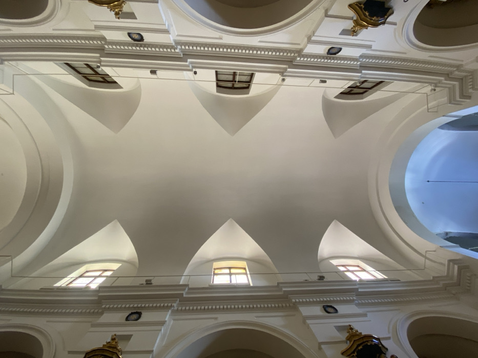 This image depicts an upward view into the grand interior architecture of the Basílica Nuestra Señora de la Merced (Basilica of Our Lady of Mercy), located in the San Nicolás neighborhood of Buenos Aires, Argentina.
The scene focuses on the church's high ceiling and vaulting, characterized by predominantly off-white, painted surfaces. A complex system of shallow, curved vaults and pendentives supports an unseen central dome structure. Four distinct, pointed, concave ceiling sections converge towards the center of the frame. Six rectangular windows with dark, possibly wooden, frames are visible, set into curved recesses at the base of the main vaulting, admitting natural light.
Ornate white architectural elements, including fluted pilasters and decorative moldings, frame the vertical structures leading up to the vaulting. These elements feature small dark circular details, possibly medallions or rosettes, at regular intervals. Golden, intricately detailed religious ornaments or decorative features are partially visible on the lower left and right margins of the image, suggesting the presence of altars or side chapel decorations below the frame.
At the very bottom, large, rounded arches, also painted white, are visible. The arch directly below leads into a brighter, light-blue-tinted area, with a thin, dark, rod-like object hanging vertically, possibly a suspended lighting fixture or part of a chandelier. The environment is well-lit by natural light, creating soft shadows and highlighting the architectural details. No people are present or visible within the frame.