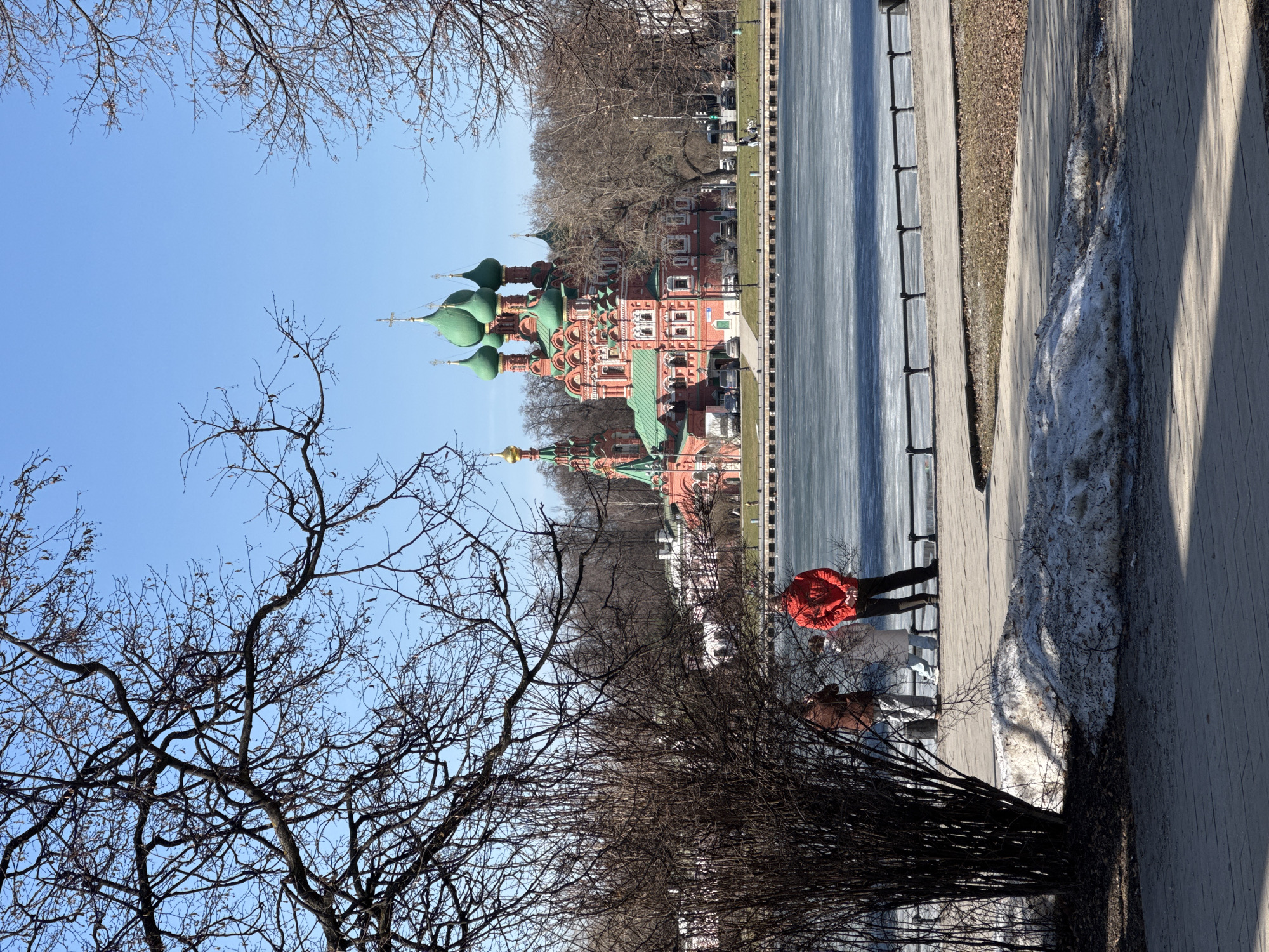 A horizontal, eye-level shot shows the Church of the Nativity of the Virgin in the city of Khimki, Russia, framed by bare trees and a partially frozen body of water. The church, constructed of red brick, is topped with green domes and gold crosses. Two people are visible on a paved walkway adjacent to the water's edge. One figure wears a red jacket and dark pants, while the second figure is clad in brown. Patches of snow are visible along the walkway.