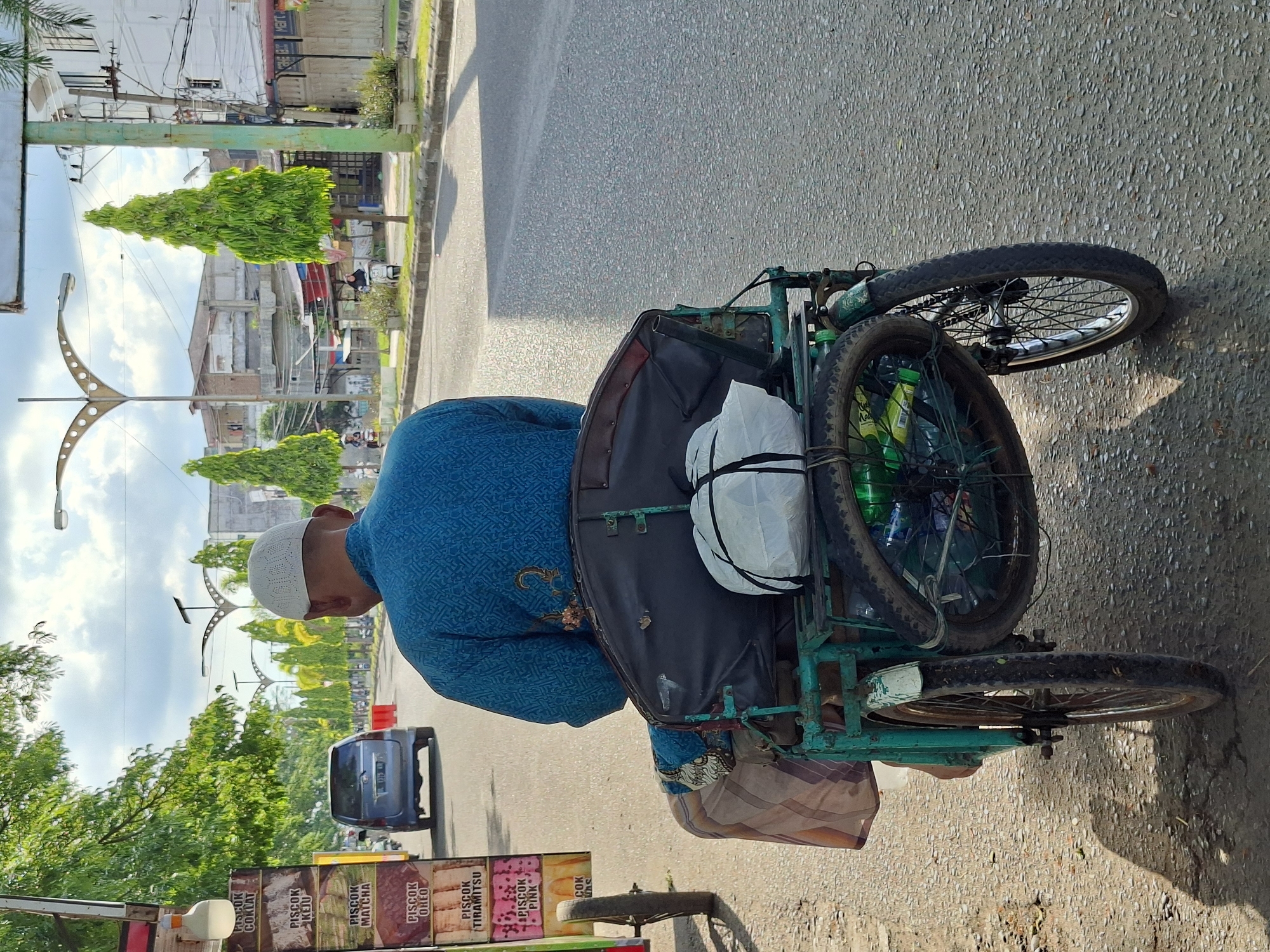 A man wearing a blue shirt and a white Muslim cap is riding a tricycle on a city street. The tricycle has three wheels and a black cargo carrier on the back. Strapped on to the cargo carrier is a white bag tied with a black rope. The man is facing away from the camera and appears to be pedaling the tricycle. In the background, there are buildings, trees, and street lights. To the man's left is a cart selling 'piscok', a street food common in Indonesia. A gray car is seen driving on the road. The location is Lhokseumawe, Indonesia.