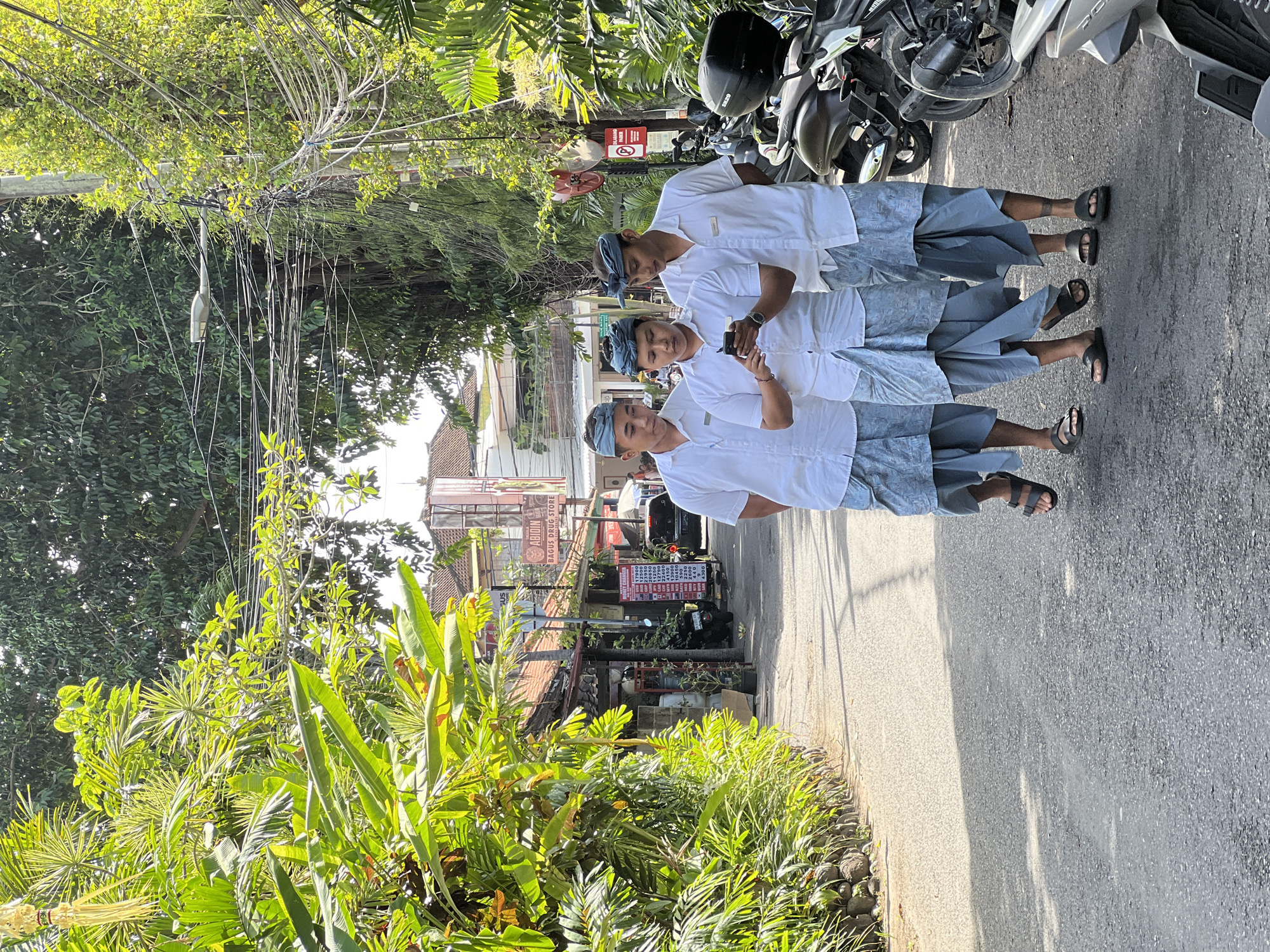 Three young men in traditional Balinese attire, consisting of white shirts and blue patterned sarongs, walk down a narrow paved street in Denpasar, Indonesia. The man in the middle is looking at a mobile phone. The street is lined with lush tropical vegetation and buildings with terracotta roofs. Several motorcycles are parked on the side of the road. Overhead, a tangle of electrical wires and a street lamp are visible amongst the trees. The lighting suggests it is daytime.