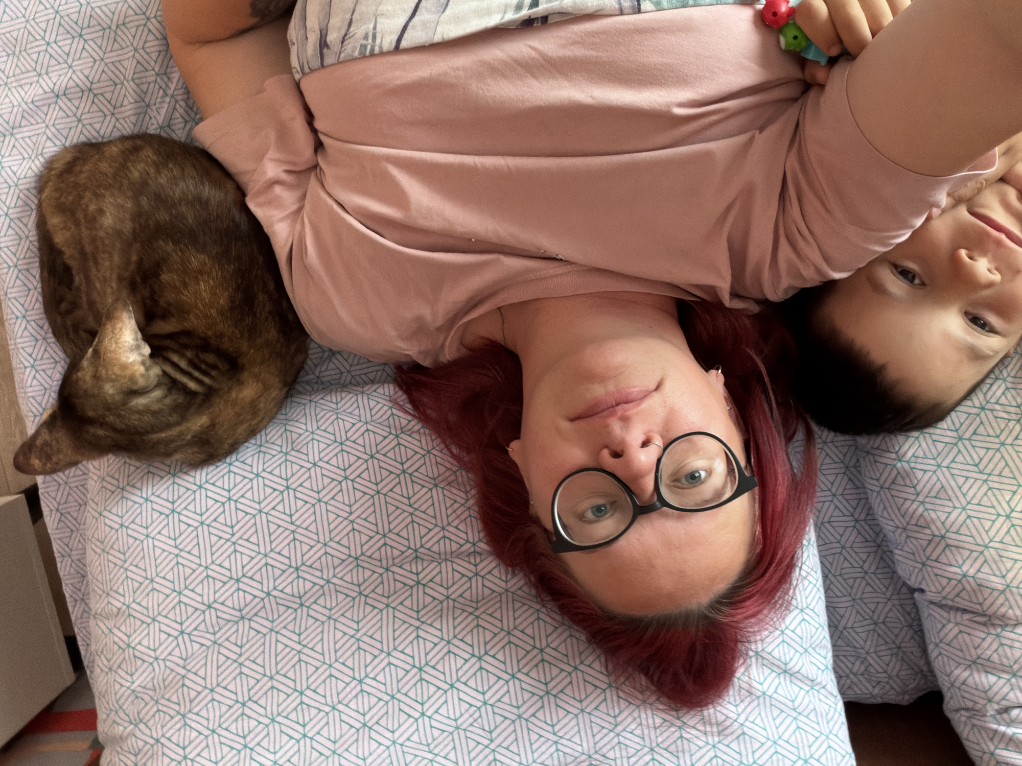 The image shows a top-down selfie of a woman and a child, likely indoors in a residential setting. The woman has red hair and is wearing glasses. A child is next to her on the right side of the frame. The child is holding a small toy. A curled-up cat is lying near the woman on the left. They are lying on a bed with a light-colored patterned bedsheet. There is a bedside table in the background. The location is presumed to be in a domestic environment, possibly in Chaikovsky, Russia.