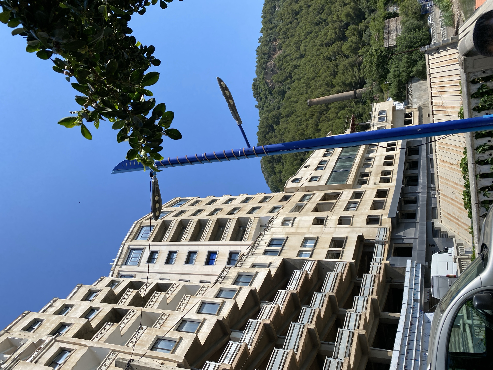 An outdoor scene captured on a bright, sunny day, featuring a distinctive building, a natural landscape, and urban infrastructure under a clear blue sky.
A multi-story building with a stepped or terraced facade dominates the right side of the frame. Its architecture features numerous recessed windows and balconies, giving it a layered appearance. The building is constructed from light-colored material, possibly concrete or stone. Some sections suggest ongoing construction or renovation, with exposed structural elements and stacks of long metal beams visible at the lower levels. A vertical garden or planted wall is visible on the far right edge of the building.
In the background, to the left of the building, a large, densely forested mountain or hillside rises steeply, covered in lush green trees. An industrial chimney or large pipe emerges from the lower slopes of this mountain, with other smaller structures nestled among the foliage.
Cutting diagonally across the mid-ground is a prominent blue pole, wrapped with copper-colored coils. Two modern, sleek street light fixtures with multiple LED lights are attached to this pole.
In the upper left corner, a tree branch laden with dark green leaves and small, round berries extends into the frame against a vibrant, cloudless blue sky.
At the bottom right, the front portions of a silver car and a white van are visible, indicating a street or parking area adjacent to the building. The scene suggests a clear, sunny day, likely around midday or early afternoon, given the bright illumination and strong shadows. There is no human activity or legible text visible.