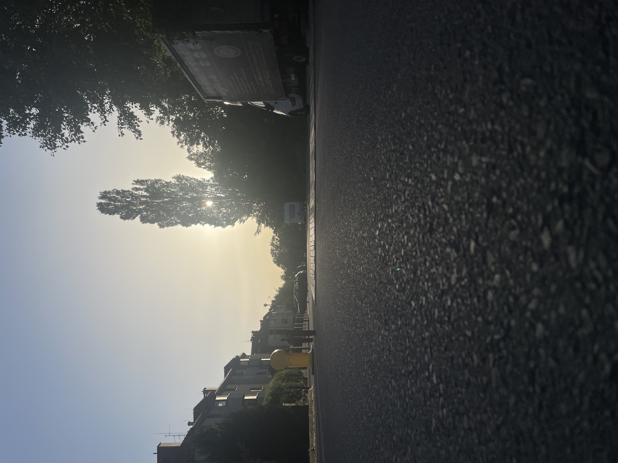 The image depicts a street scene in Wrocław, Poland. The foreground is dominated by a dark asphalt road, showing wear and tear. To the left, there is a sidewalk with yellow bollards and buildings of various heights and architectural styles. The sky is bright with the sun obscured by trees, creating a hazy atmosphere. Along the right side of the street, there are trees and parked vehicles. A person is partially visible walking along the street.
