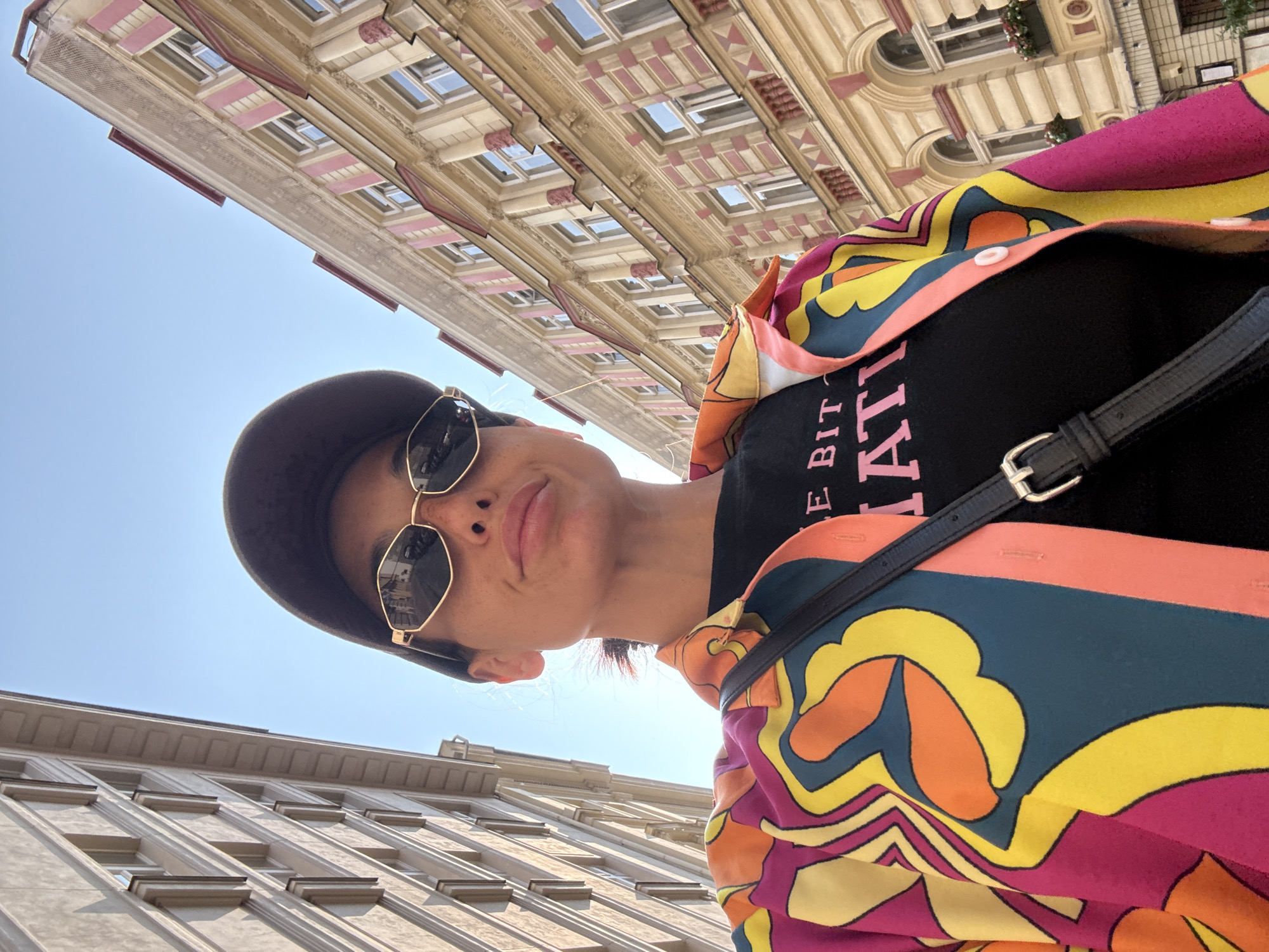 A low-angle, full-body outdoor selfie is taken on a street in Prague, Czechia, with a woman wearing a colorful shirt, black t-shirt, black baseball cap, and gold-rimmed sunglasses. The woman is positioned in the center of the frame, facing the camera. Her black t-shirt has pink text that reads "E BIT AT". She wears a black crossbody bag. The background consists of two buildings on either side of the street and a clear blue sky. The building on the left appears to be an apartment building with beige facades and numerous windows. The building on the right is a yellow building with pink accents and arched windows, adorned with flowers in window boxes. Sunlight illuminates the scene, creating shadows on the buildings and the woman's face.