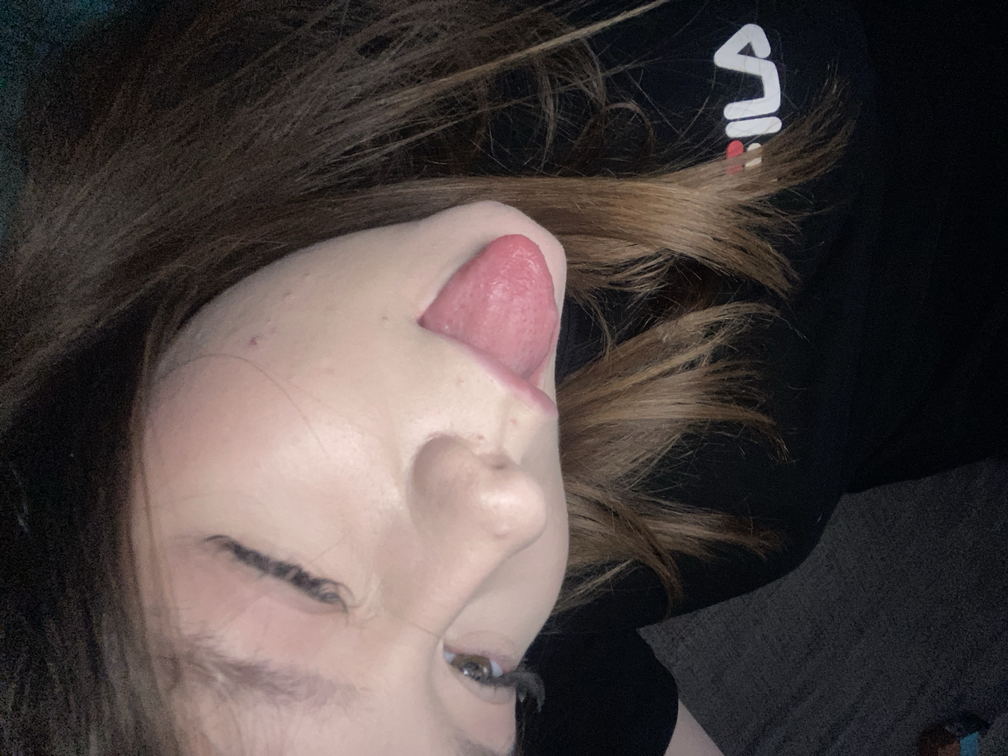 A close-up, tilted shot shows a person with light skin, dark brown hair, and one eye visible. They are sticking their tongue out. The person is wearing a black shirt with a white "FILA" logo on the right side. The background consists of dark colors, possibly a couch or another surface. No evidence suggests the location is Chaikovsky, Russia.