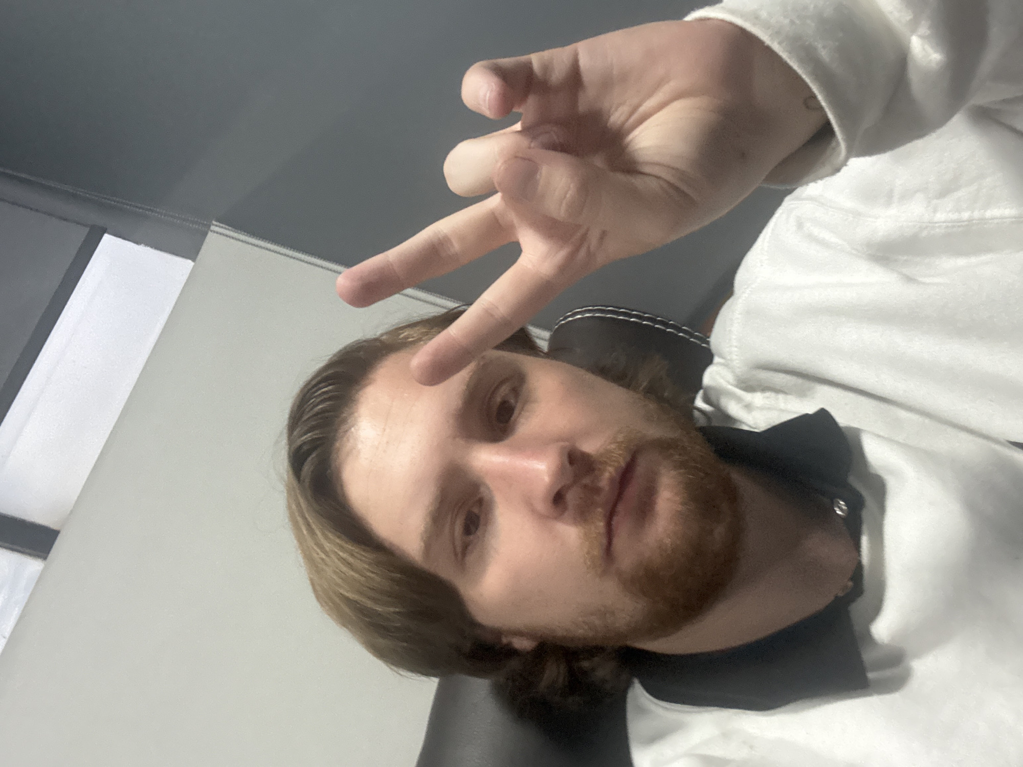 A selfie of a fair-skinned man with medium-length brown hair and a beard, wearing a white hoodie. His right hand is raised, making a "W" gesture. The background includes a gray wall and what appears to be a window with light filtering through. The setting is possibly indoors. There is no specific information available to pinpoint the location to Cuautitlán Izcalli, Mexico.