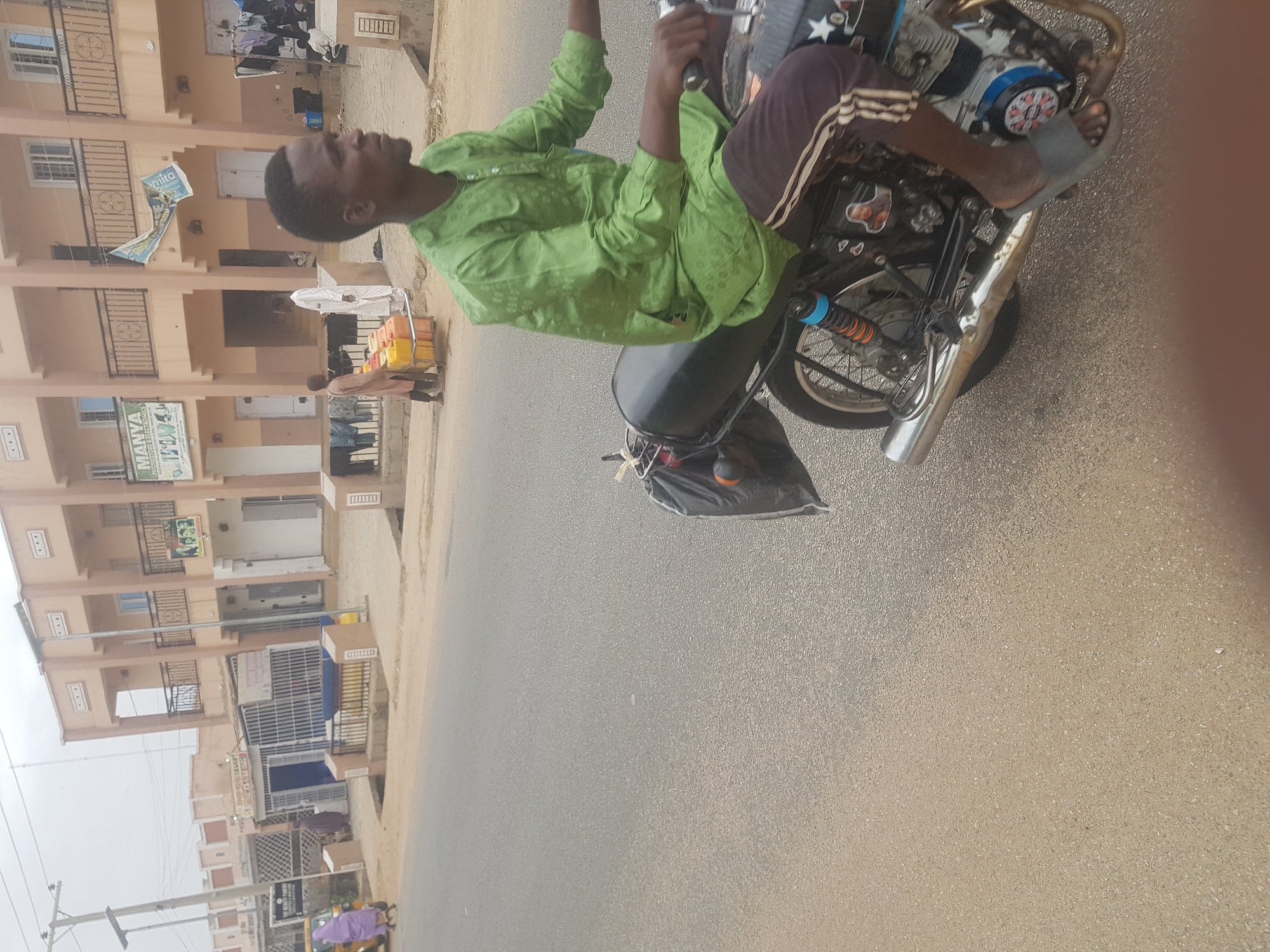 A man rides a motorcycle on a street in Dabawa, Nigeria. The man, dressed in a green shirt and brown shorts, sits astride the motorcycle, which is equipped with a rear rack. He is wearing a flip-flop on his right foot. The road is paved and beige. The background shows multi-story buildings with balconies. There is a sign on one of the buildings with the name "Manya Pharmacy". A pedestrian is visible near the pharmacy, pulling a cart. Further down the street, a purple tuk-tuk vehicle is parked.