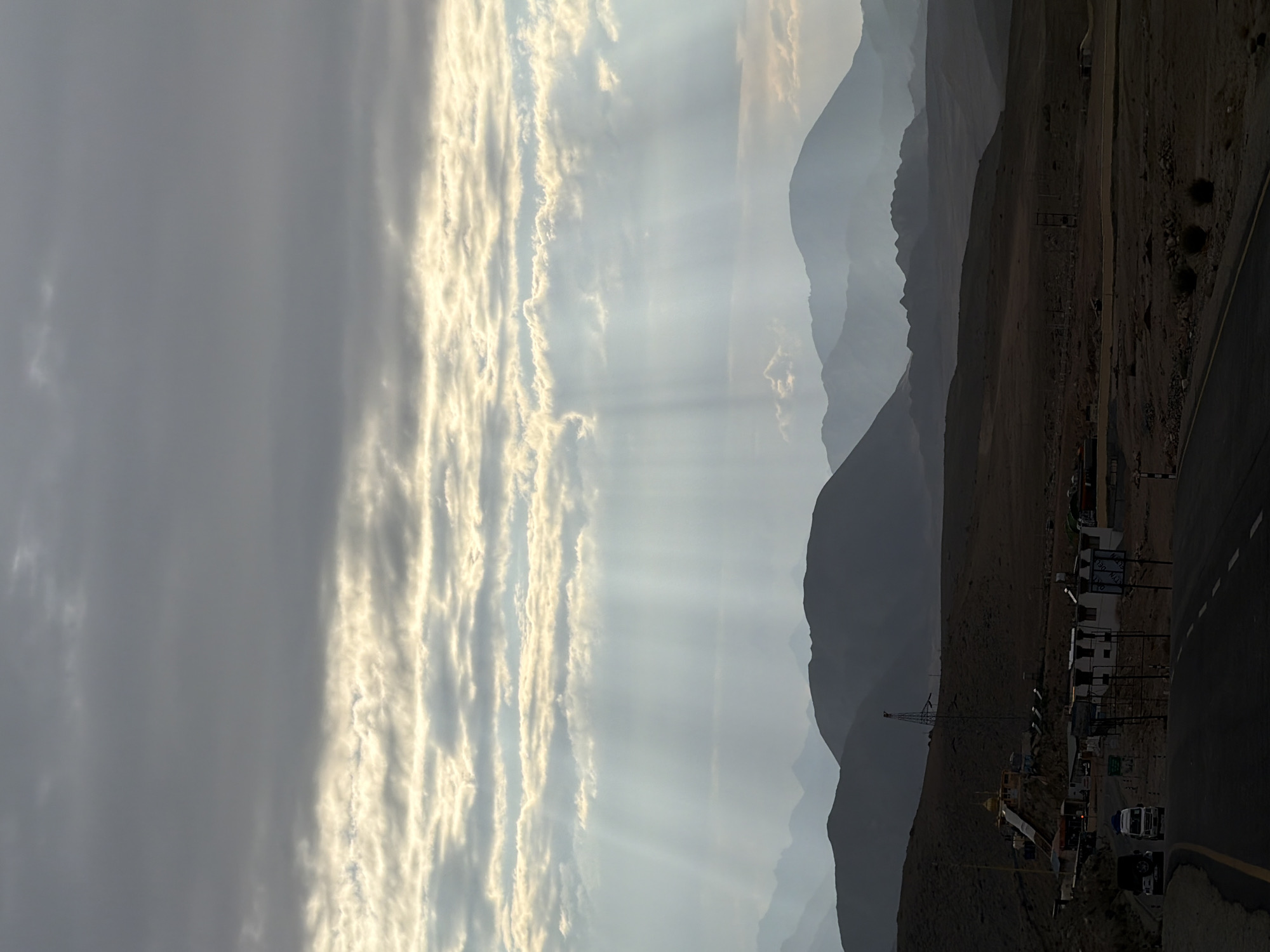 The image depicts a dramatic mountainous landscape at dusk, with the sun's rays piercing through a cloudy sky and casting ethereal beams onto the terrain. In the foreground, a paved road curves to the right, with two vehicles, a white van and a dark SUV, traveling away from the viewer. A small settlement is visible on the left side of the road, featuring several low-rise buildings, a religious structure with a golden dome, and a tall tower with antennae. The settlement appears to be located in a dusty, arid environment. The middle ground is characterized by rolling hills and rugged terrain, leading to a series of majestic, layered mountains in the background that fade into the hazy sky. The overall atmosphere is serene and majestic, with a sense of remoteness and natural grandeur. The image appears to be taken in Chumkar, India, a region known for its stunning mountain scenery.
