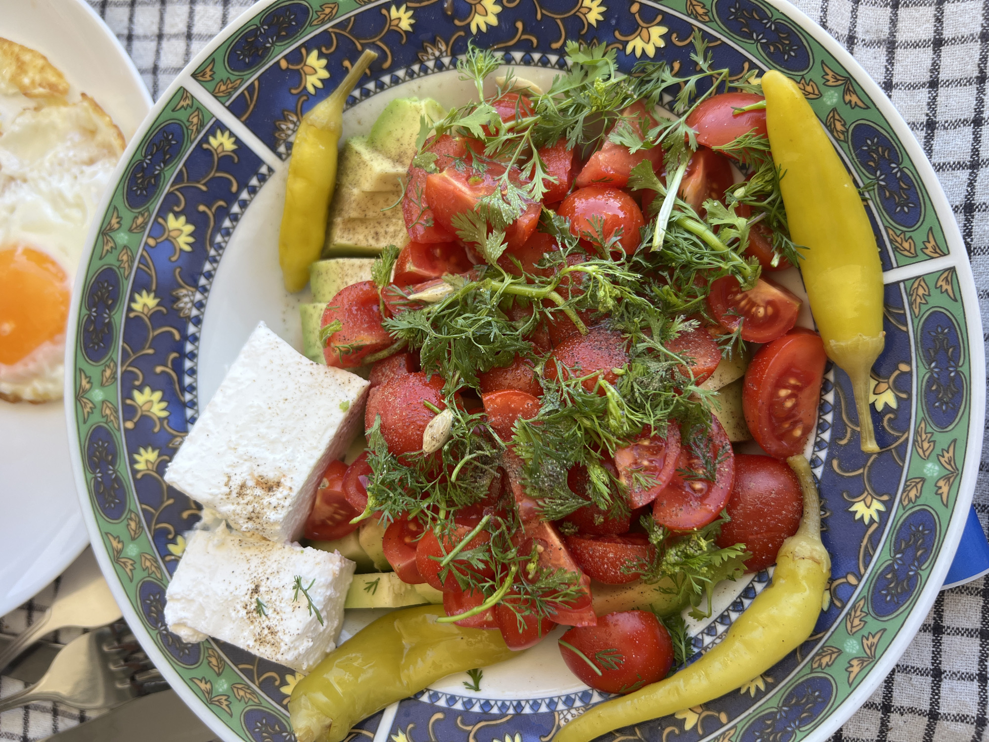 In Baošići, Montenegro, a close-up shot captures a plate of food on a table with a checkered tablecloth. The plate, decorated with a blue, green, and gold pattern, holds a salad composed of chopped tomatoes, sliced avocado, and fresh dill. Two large blocks of white cheese, sprinkled with seasoning, are placed on top. Several whole, yellow pickled peppers are arranged around the salad, with two visible on the left and two on the right side of the plate. In the background, a portion of a white plate with a fried egg is visible. Utensils, likely forks, are also present in the upper left corner, partially obscured. The lighting suggests an outdoor setting.