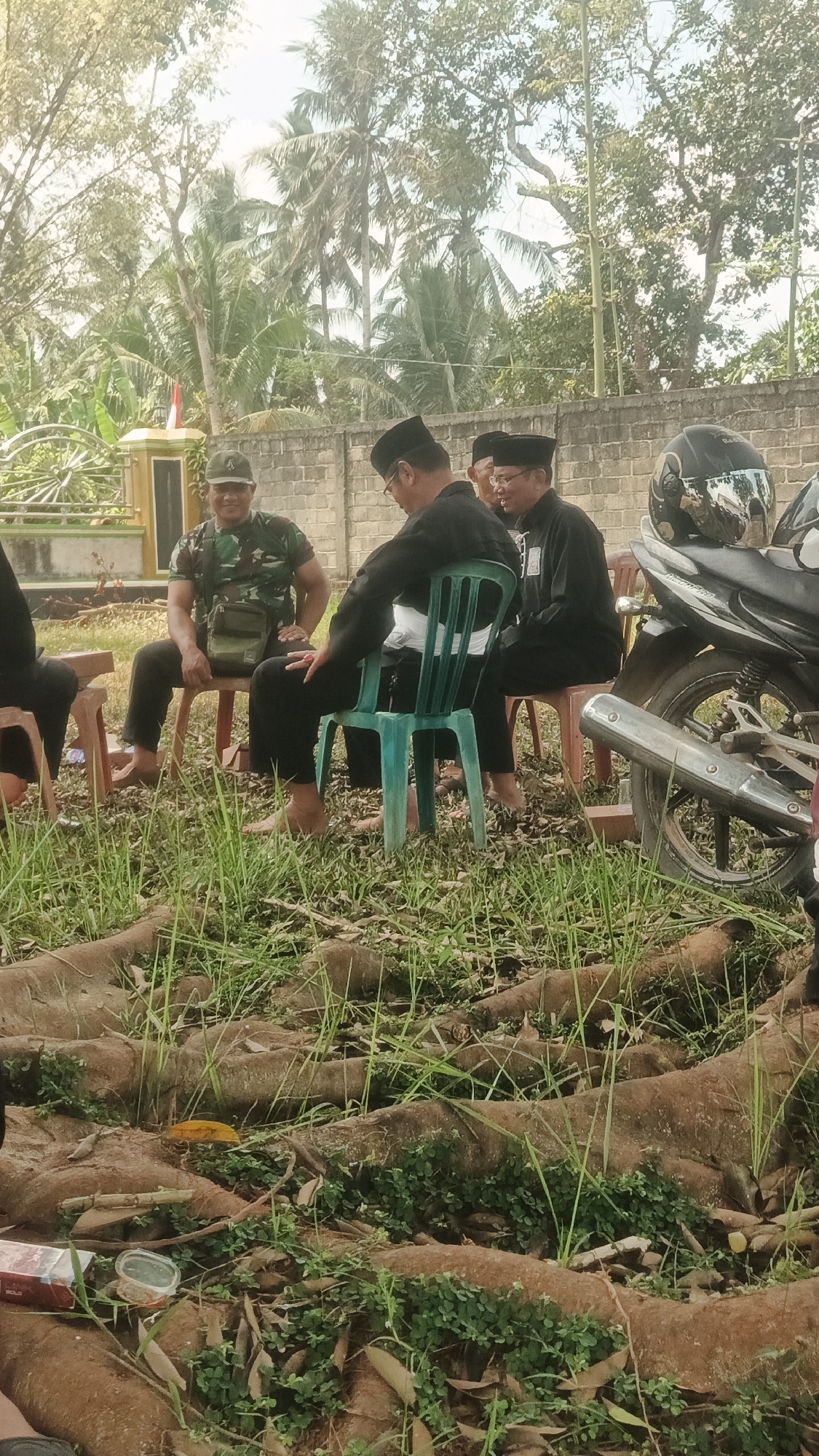 In Nabang Baru, Indonesia, three men are seated outdoors, appearing to be in a casual gathering. One man, dressed in military camouflage, sits on a brown chair. To his right, a man in a black shirt and peci (a type of cap) is seated on a turquoise chair, facing left. Another man, also in a black shirt and peci, is seated on a red chair to the right of the man on the turquoise chair, and is partially obscured. The men are engaged in conversation. The environment is natural, with lush green grass and exposed tree roots in the foreground, and trees and a concrete wall in the background. A motorcycle is parked to the right of the frame.