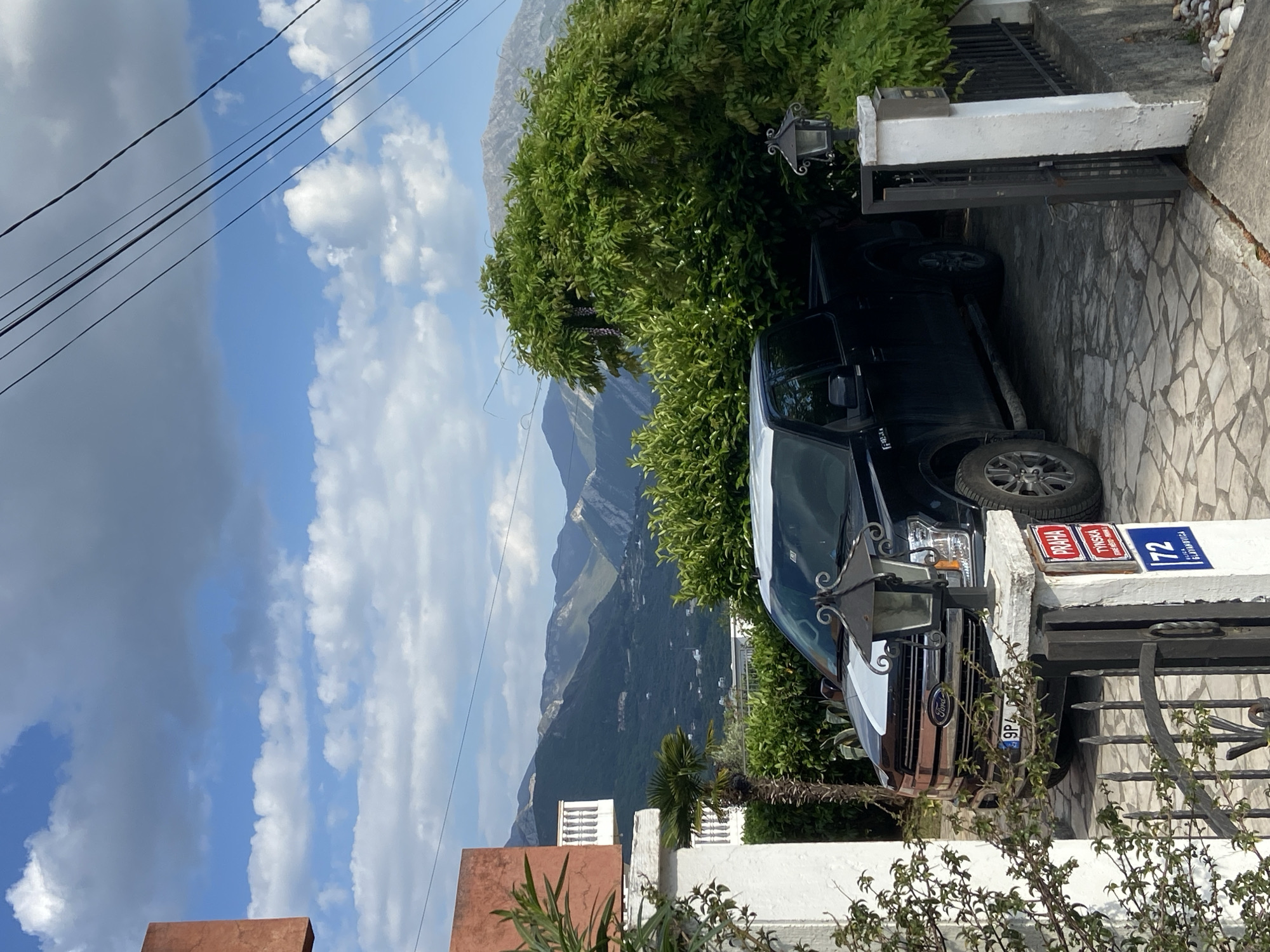The image shows a black Ford F-150 parked in a driveway in the city of Bar, Montenegro. The vehicle is partially obscured by foliage and a gate. A white gatepost displays two street signs: one reading "PRAHA TIVATSKA" in red lettering and another with the number "72". Mountains are visible in the background under a partly cloudy sky. Power lines are visible in the upper left corner of the image. The driveway is paved with stone tiles. The location appears to be a residential area.